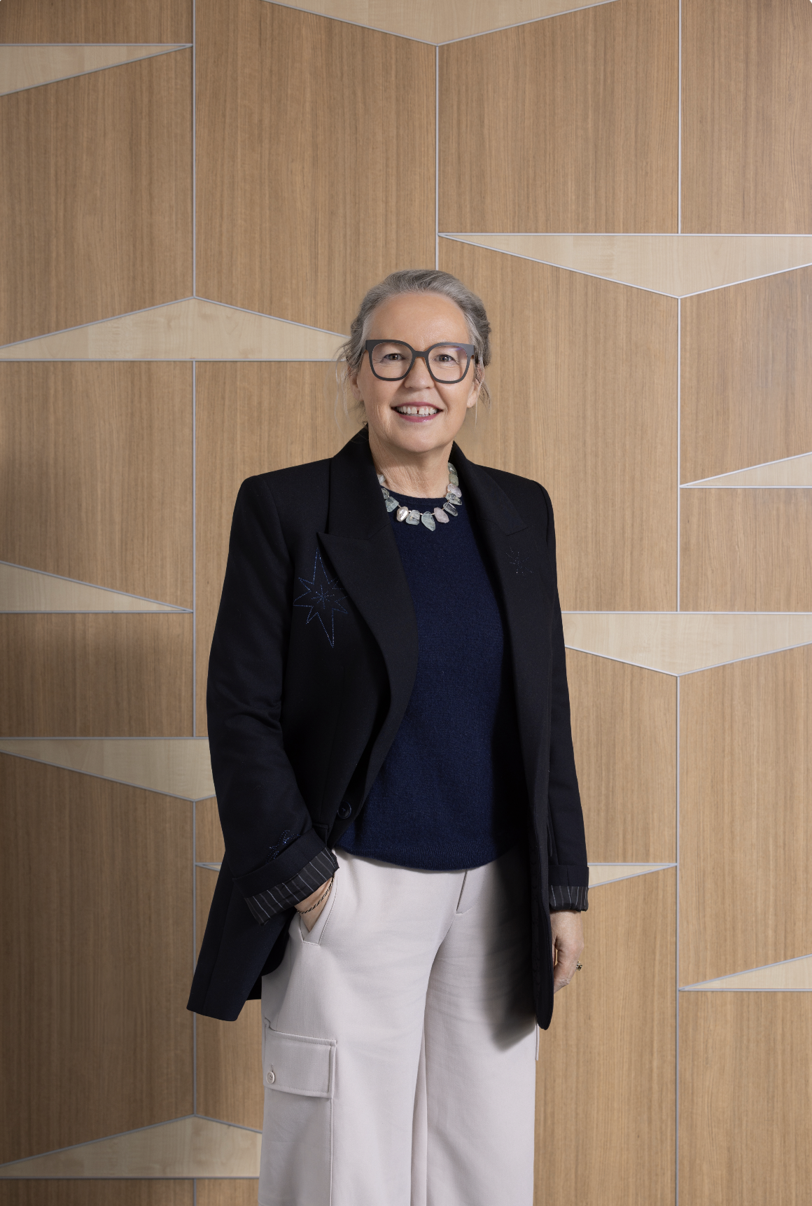 A stylish woman with gray hair and glasses stands in front of a wooden geometric-patterned wall, smiling, wearing a black blazer, navy sweater, beige pants, and a chunky necklace.