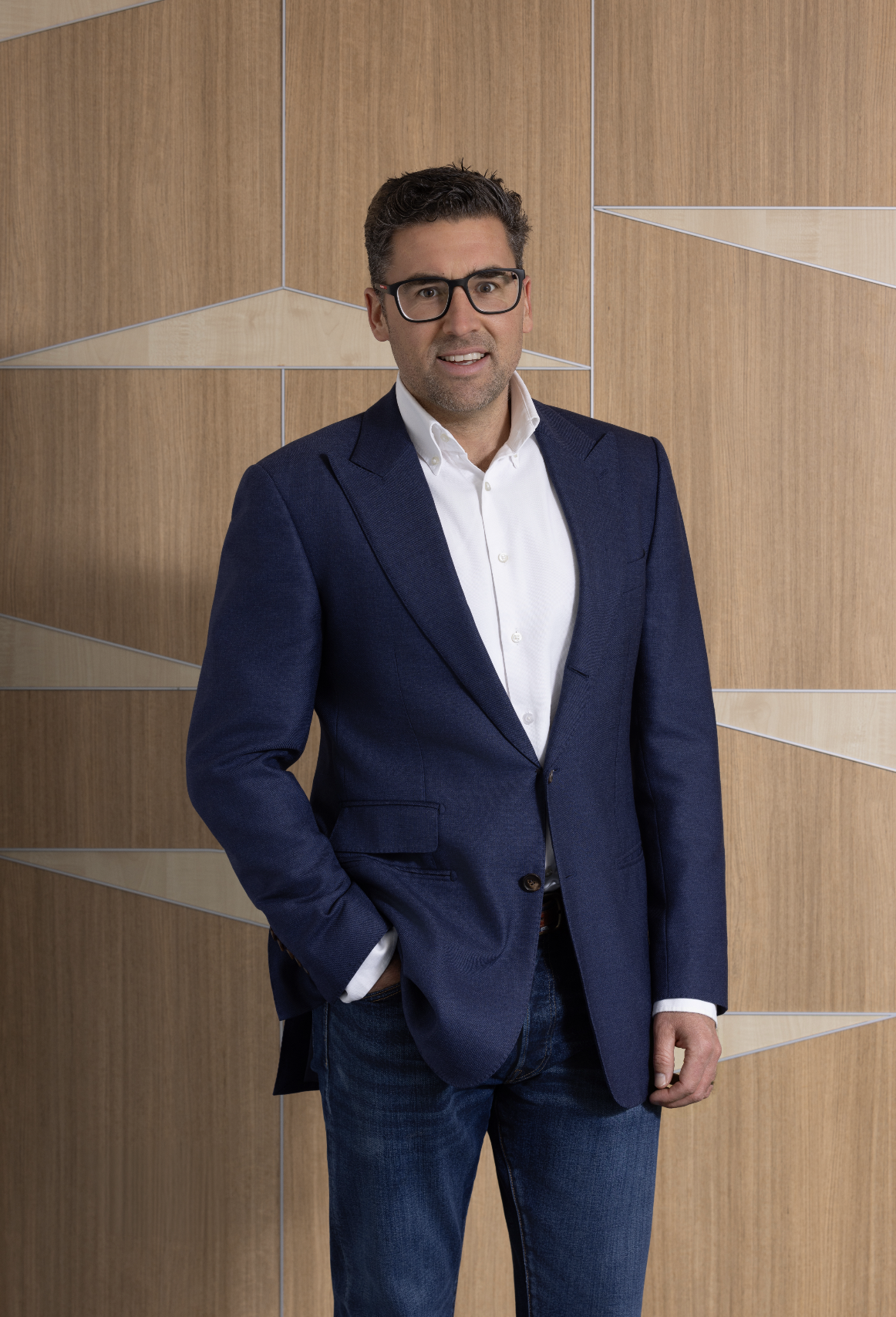 Man in a navy blazer, white shirt, and dark jeans standing in front of a wooden wall with geometric design.