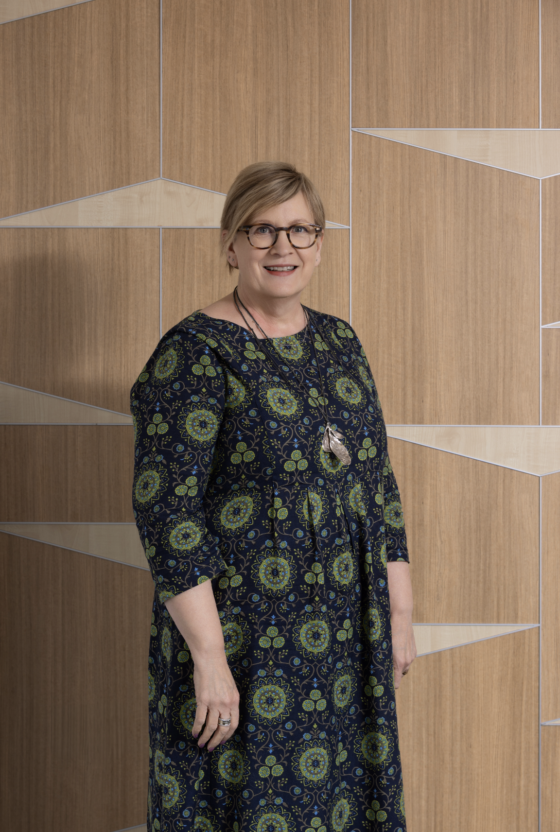 A woman with glasses and short blonde hair smiling, wearing a dress with a blue and green floral pattern, standing in front of a wooden wall with geometric paneling.