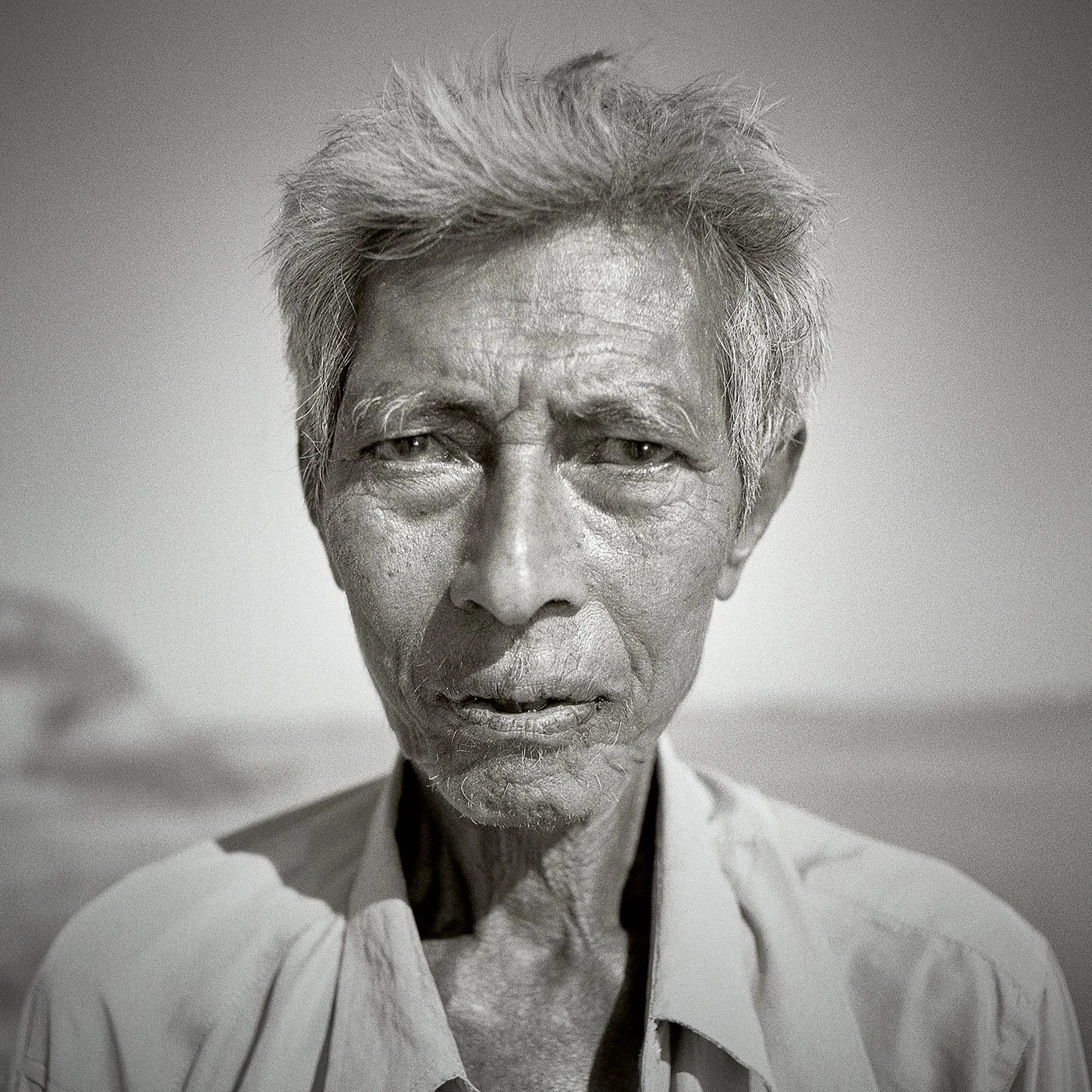 Un portrait en noir et blanc d'un homme âgé avec des cheveux gris, regards perçants, visage marqué par le temps, portant une chemise ouverte