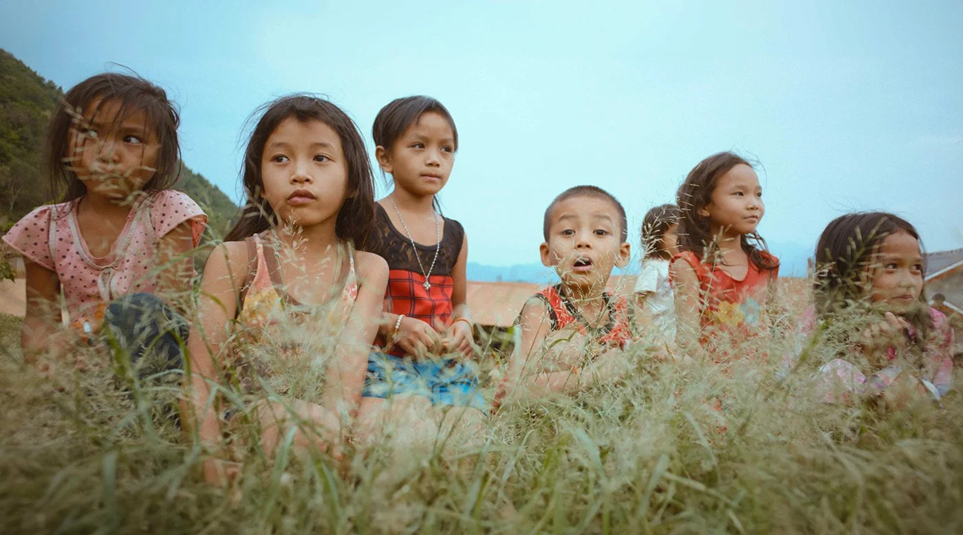 Groupe de sept enfants assis dans l'herbe, regardant vers l'objectif, avec une maison en arrière-plan et un ciel clair.