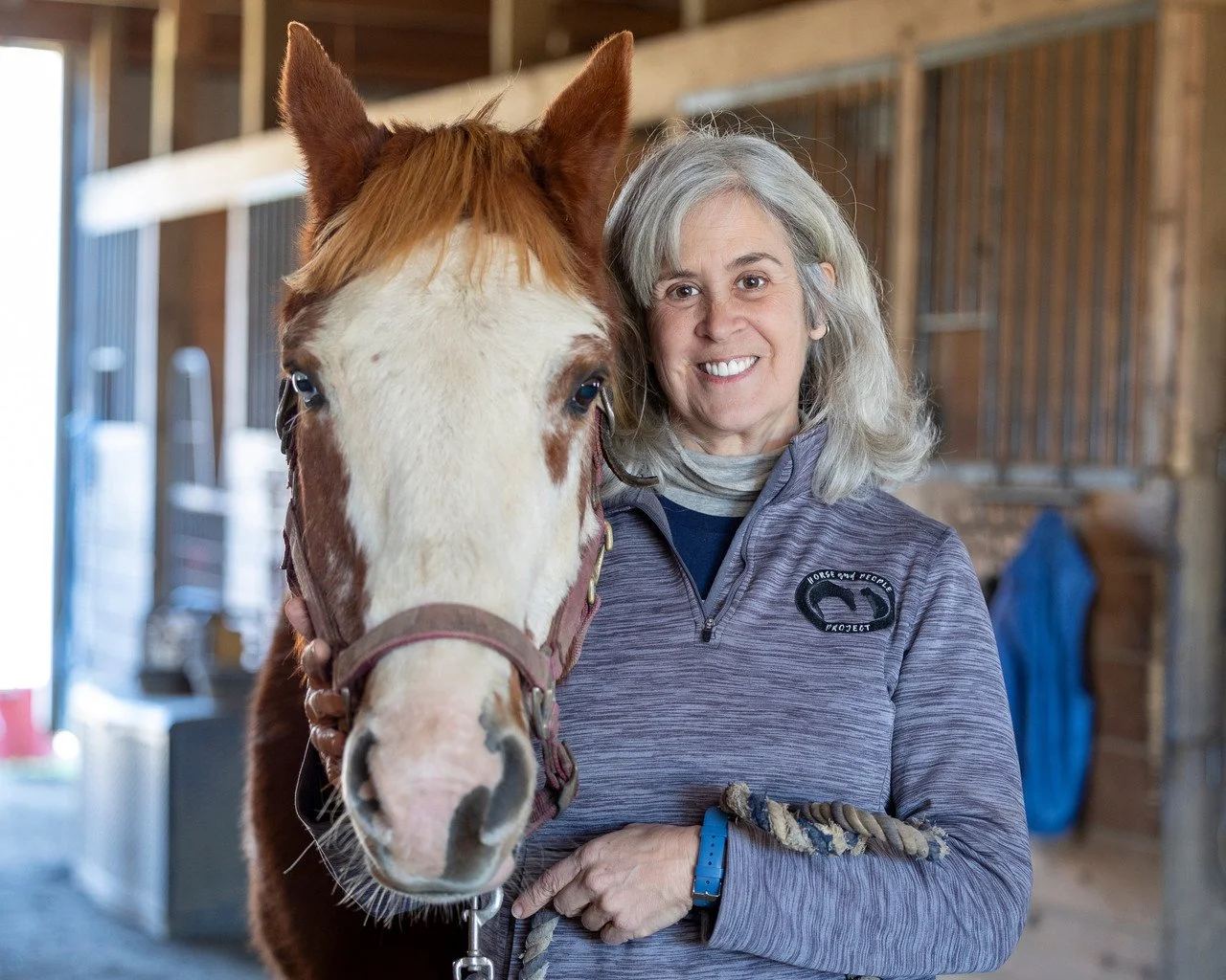 Maria Alessandri standing next to a brown and white horse inside a barn.