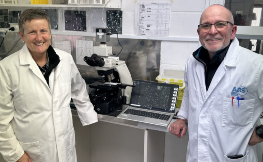 Two scientists in lab coats standing in a laboratory next to a microscope and a laptop, smiling at the camera.