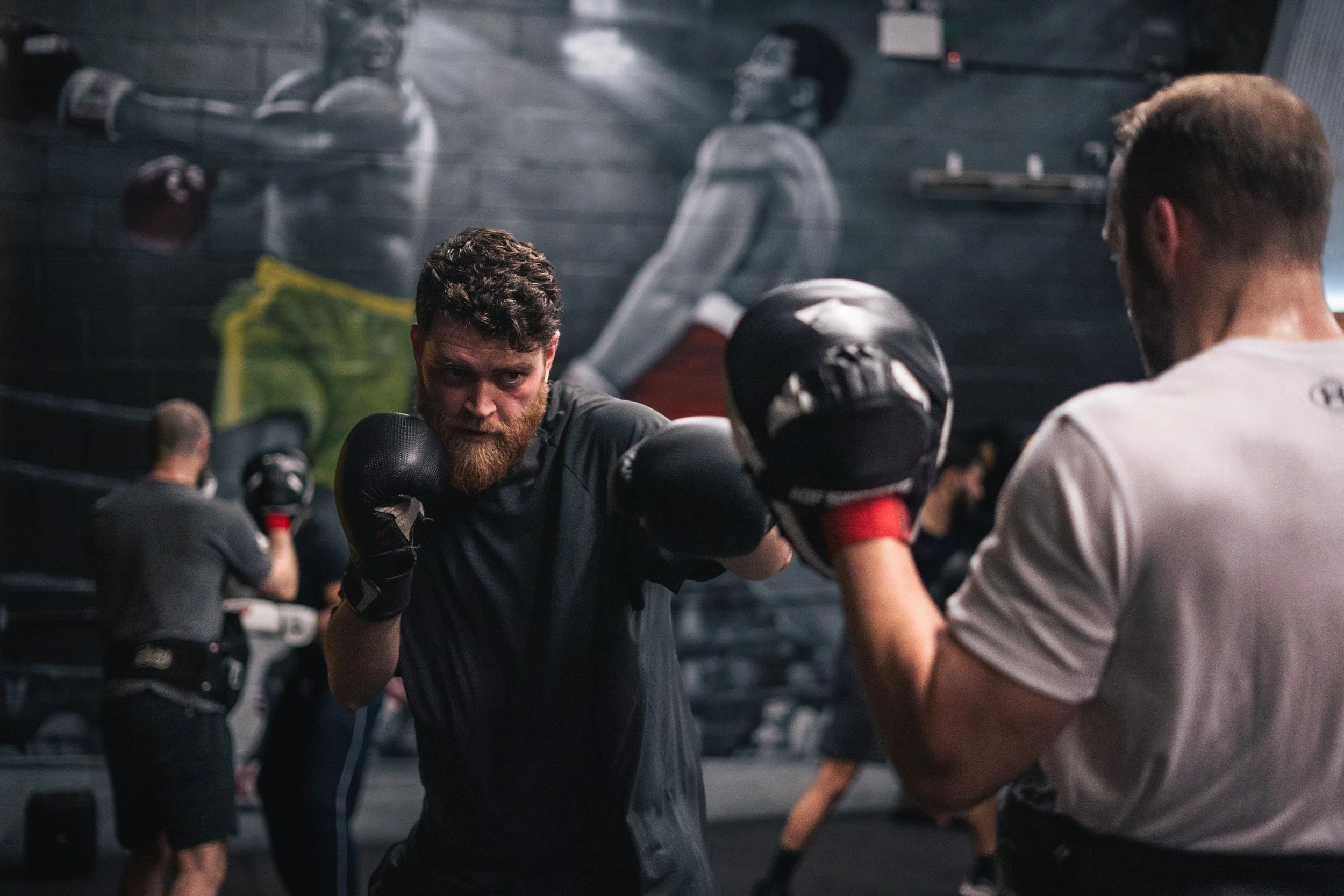 Two men practicing boxing in a gym with a dark wall mural, one wearing black gloves and the other wearing white gloves, and their backs are facing the camera.