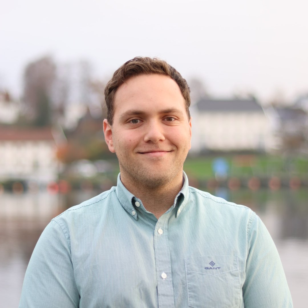 En menn står foran en vannflate med bygninger i bakgrunnen, iført lys blå skjorte og smilende.