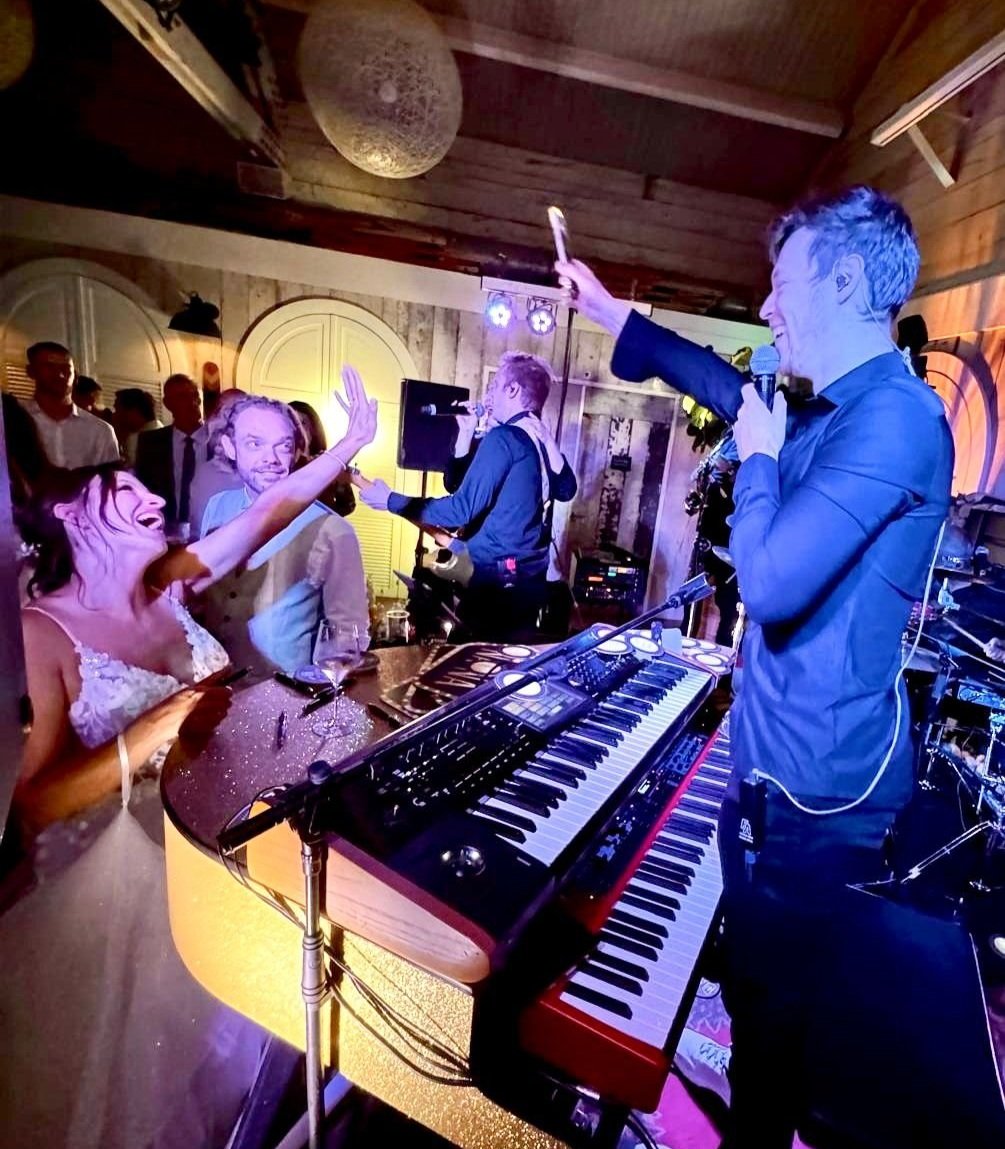 Mensen die genieten van livemuziek bij een evenement, met een vrouw in een bruidsjurk die enthousiast naar een toetsenist op het podium zwaait.