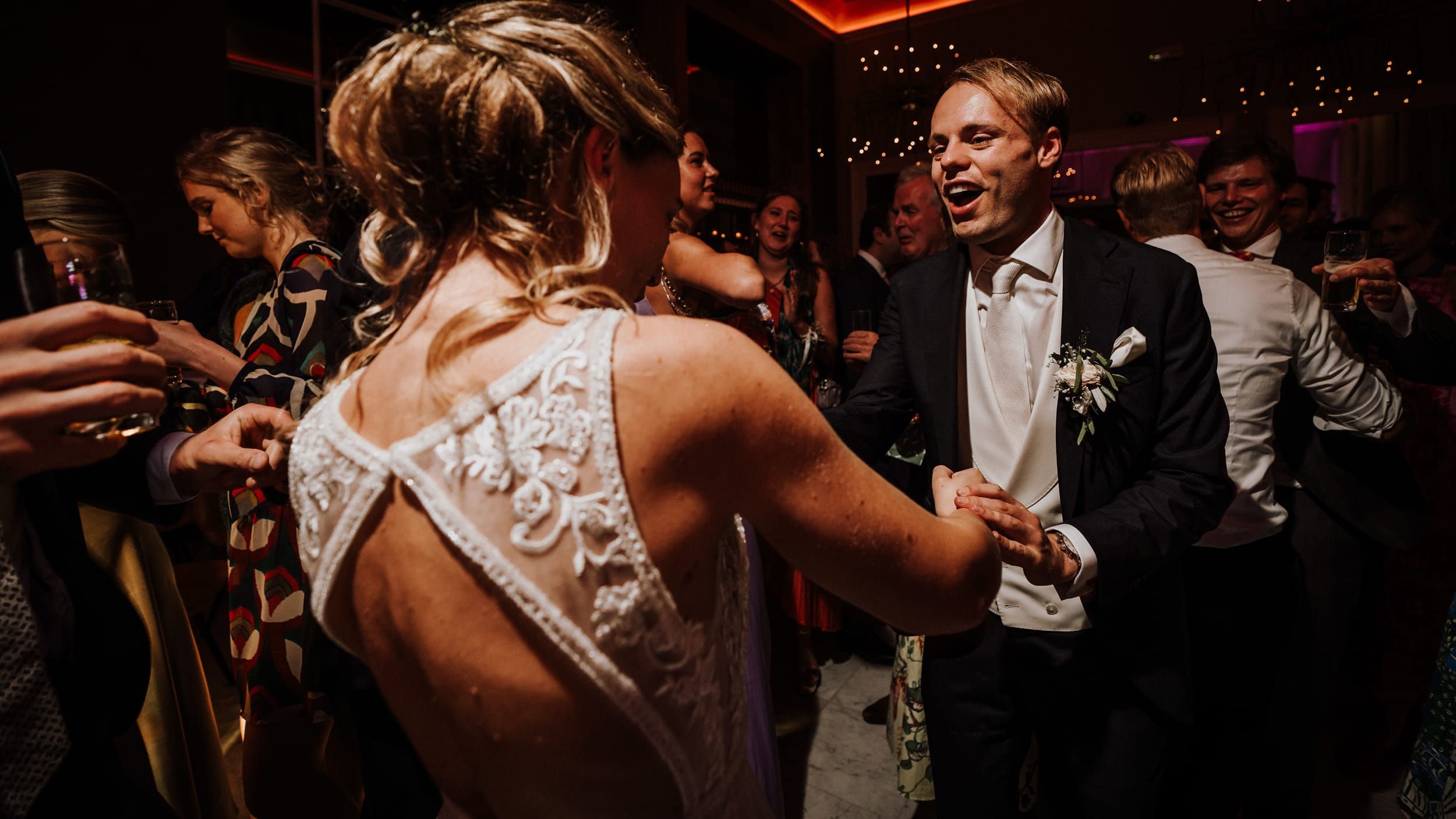 Mensen dansen op een feest, bruid in een witte jurk met kant details en bruidegom in een donker pak met bloemen op de revers.