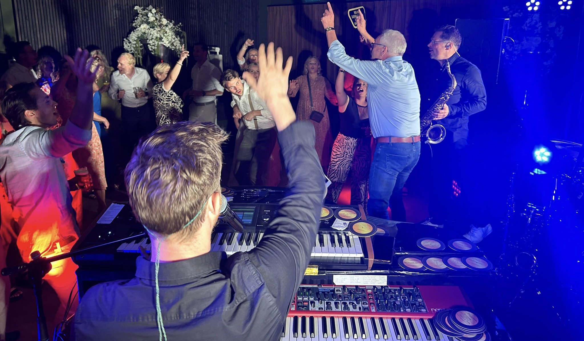 Mensen dansen en vieren feest bij een muzikant met een keyboard en een saxofonist op de achtergrond, met feestverlichting.