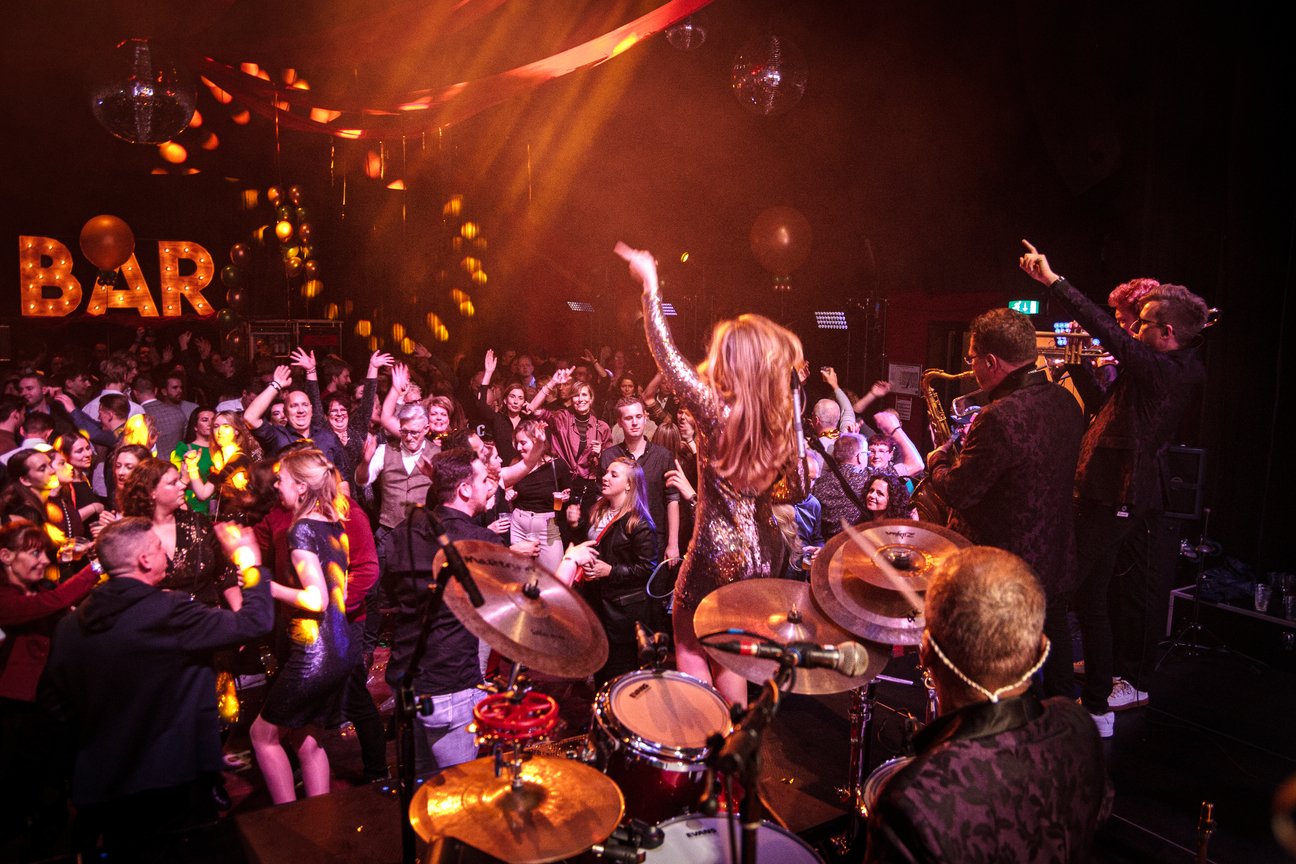 Mensen dansen op een feest met een liveband; "BAR" verlicht op de achtergrond.