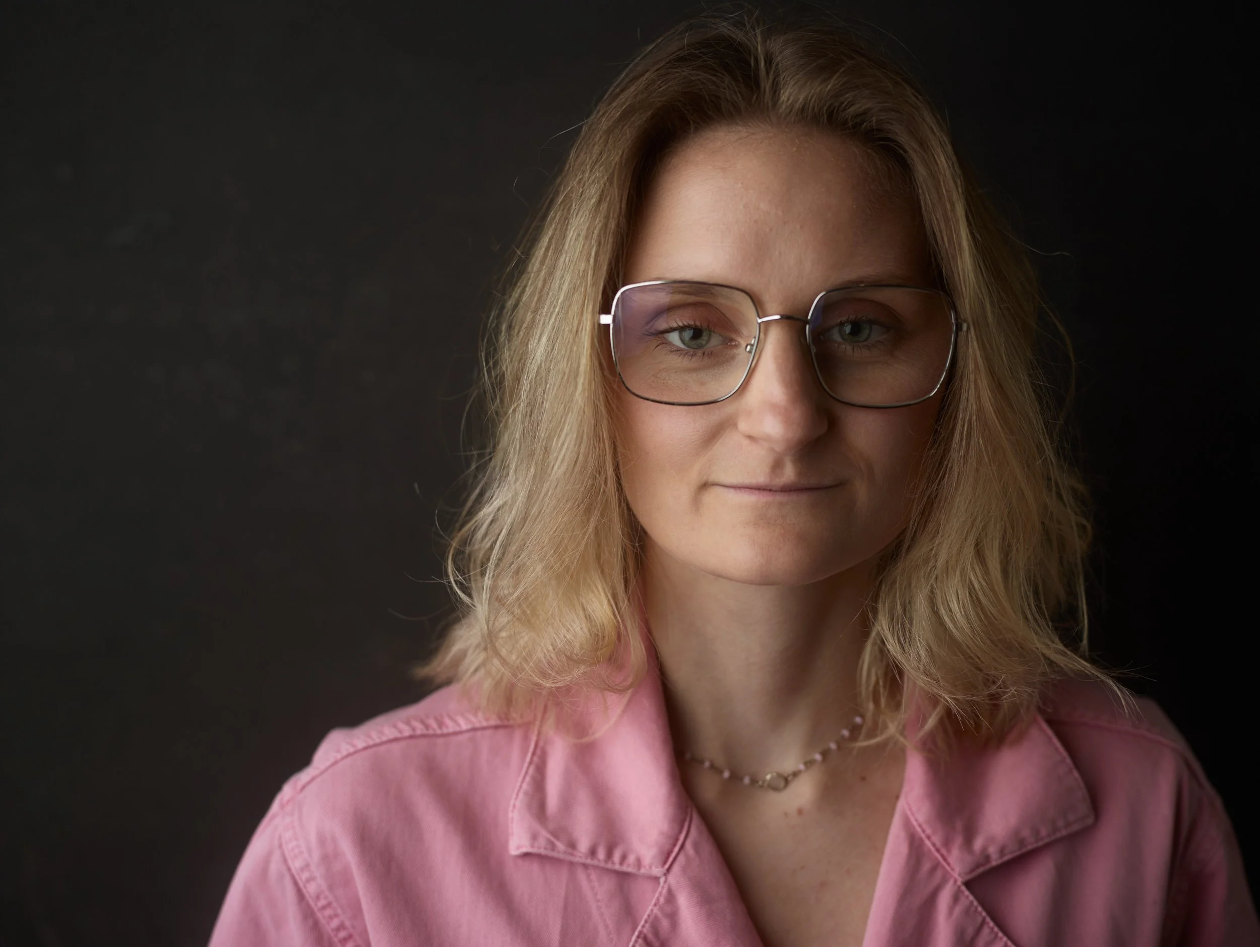 A woman with shoulder-length blonde hair wearing glasses and a pink shirt against a dark background.