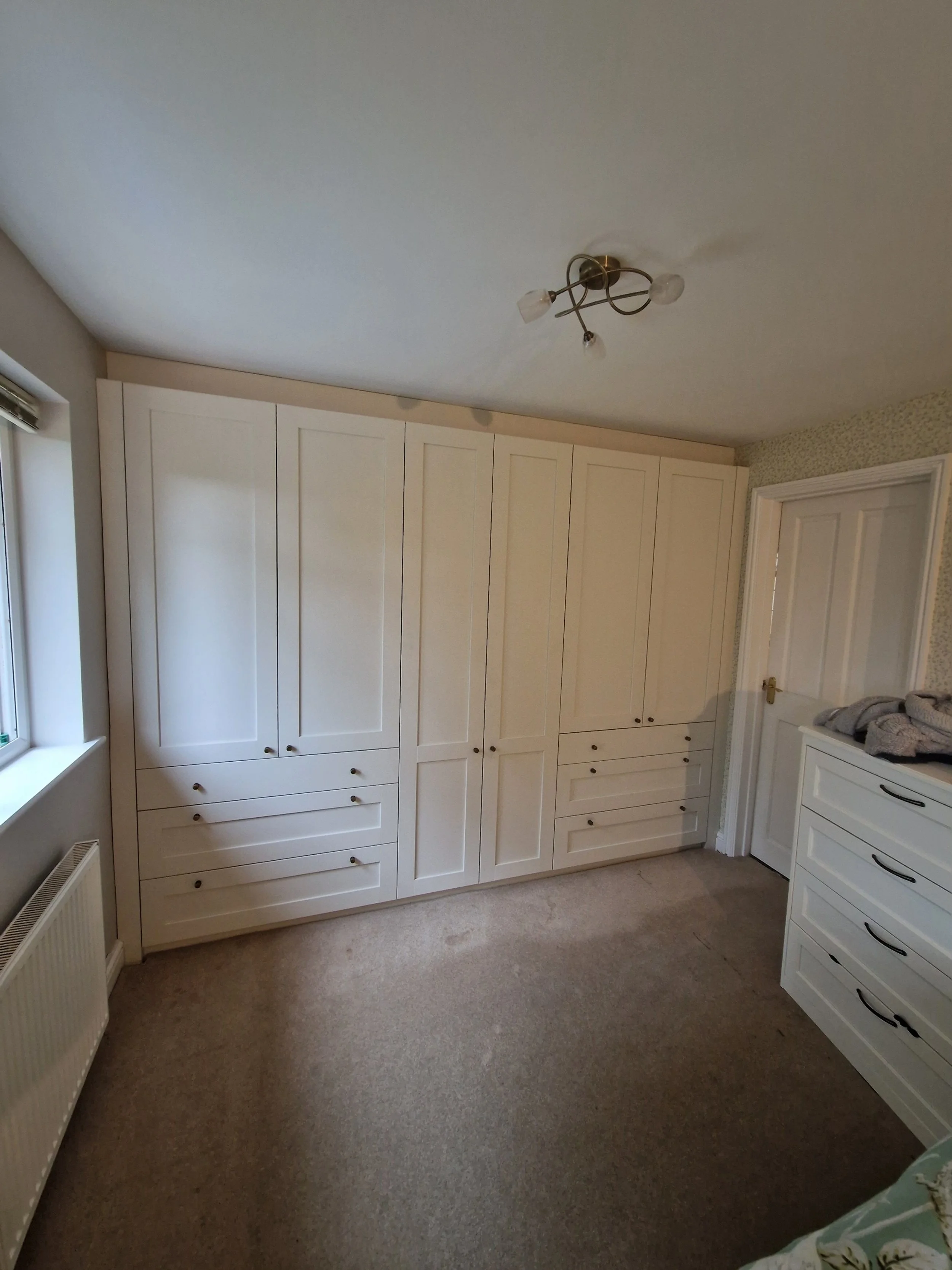 A room with beige carpeted floor, a large white wardrobe with multiple drawers and cabinets along the wall, a white door with a brass doorknob, and a window with a radiator below it. There is also a white dresser with stacked clothes on top.