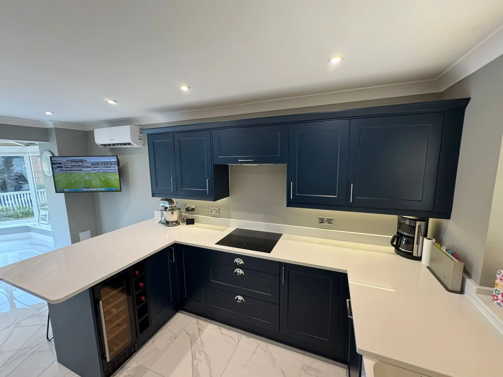 A modern kitchen with dark blue cabinets, white countertops, and marble flooring. A window with a sliding door leads to an outdoor deck, and a television is mounted on the wall. There are small appliances on the counter, and a wine rack is built into the island.