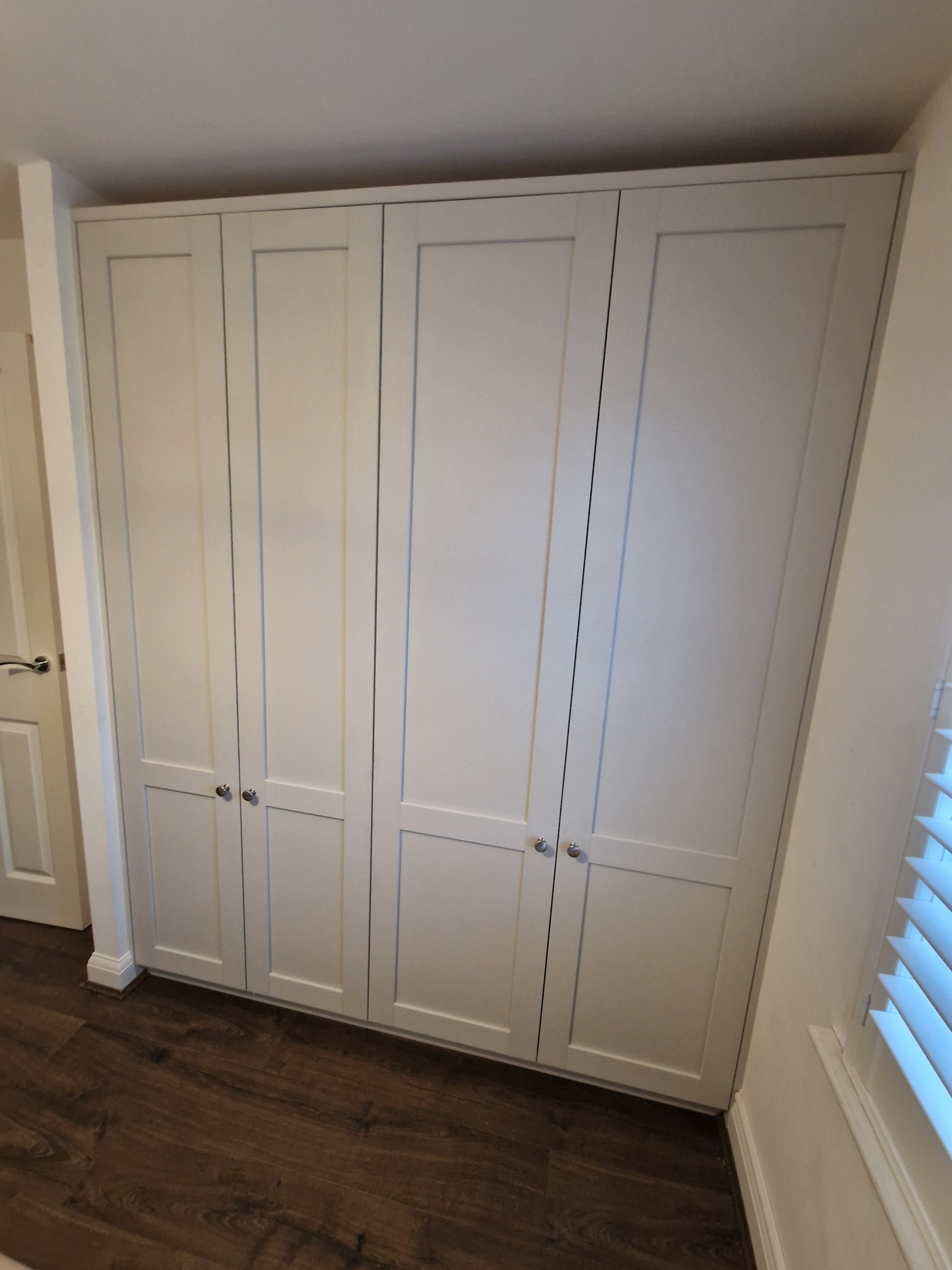 A large white built-in closet with multiple panels and round silver knobs, situated against a wall in a room with dark hardwood flooring and a window with white blinds.