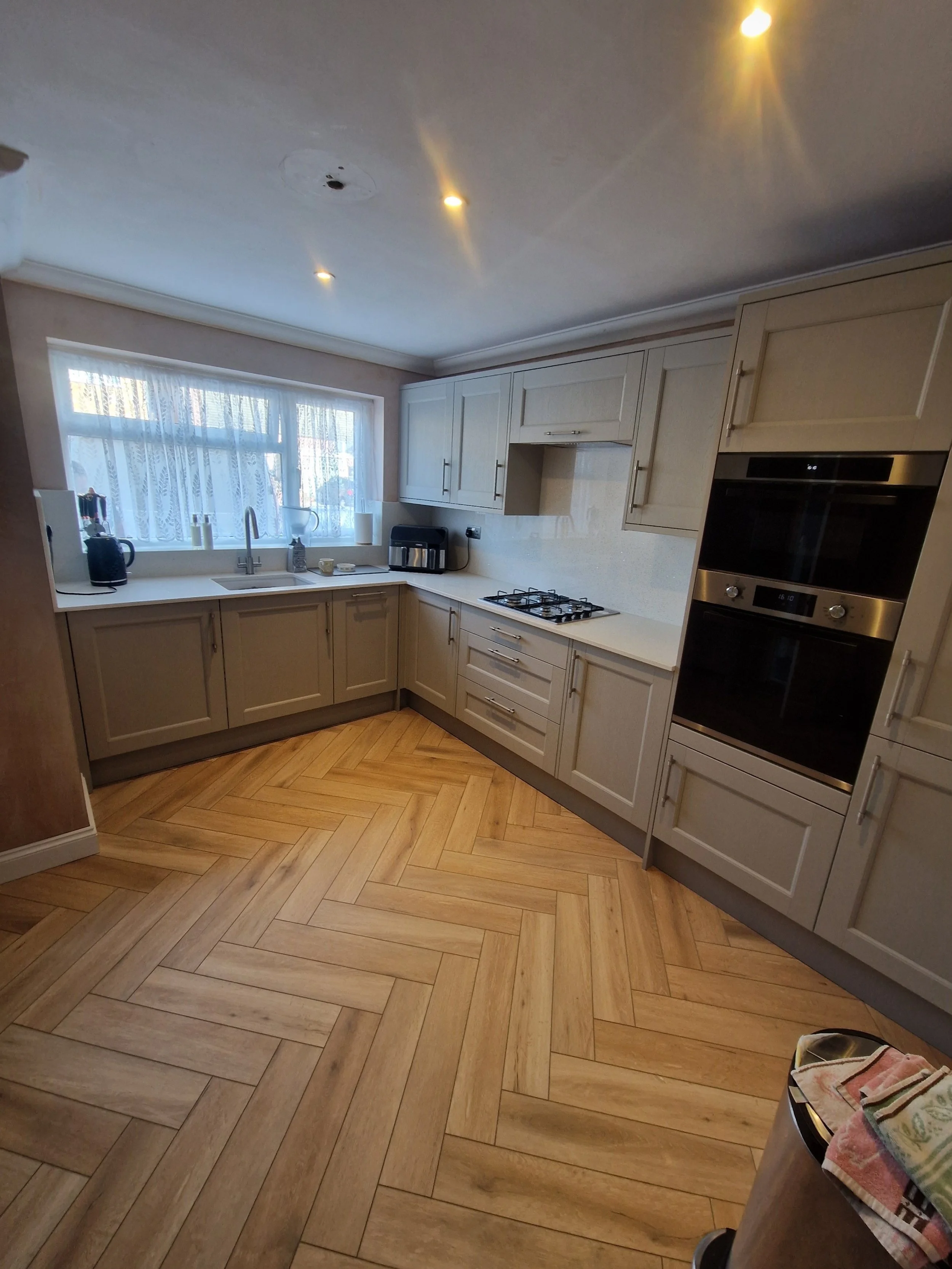 A kitchen with beige cabinets, a white countertop, a window with lace curtains, and wooden herringbone flooring. There are appliances like a microwave, oven, and kettle on the countertop.