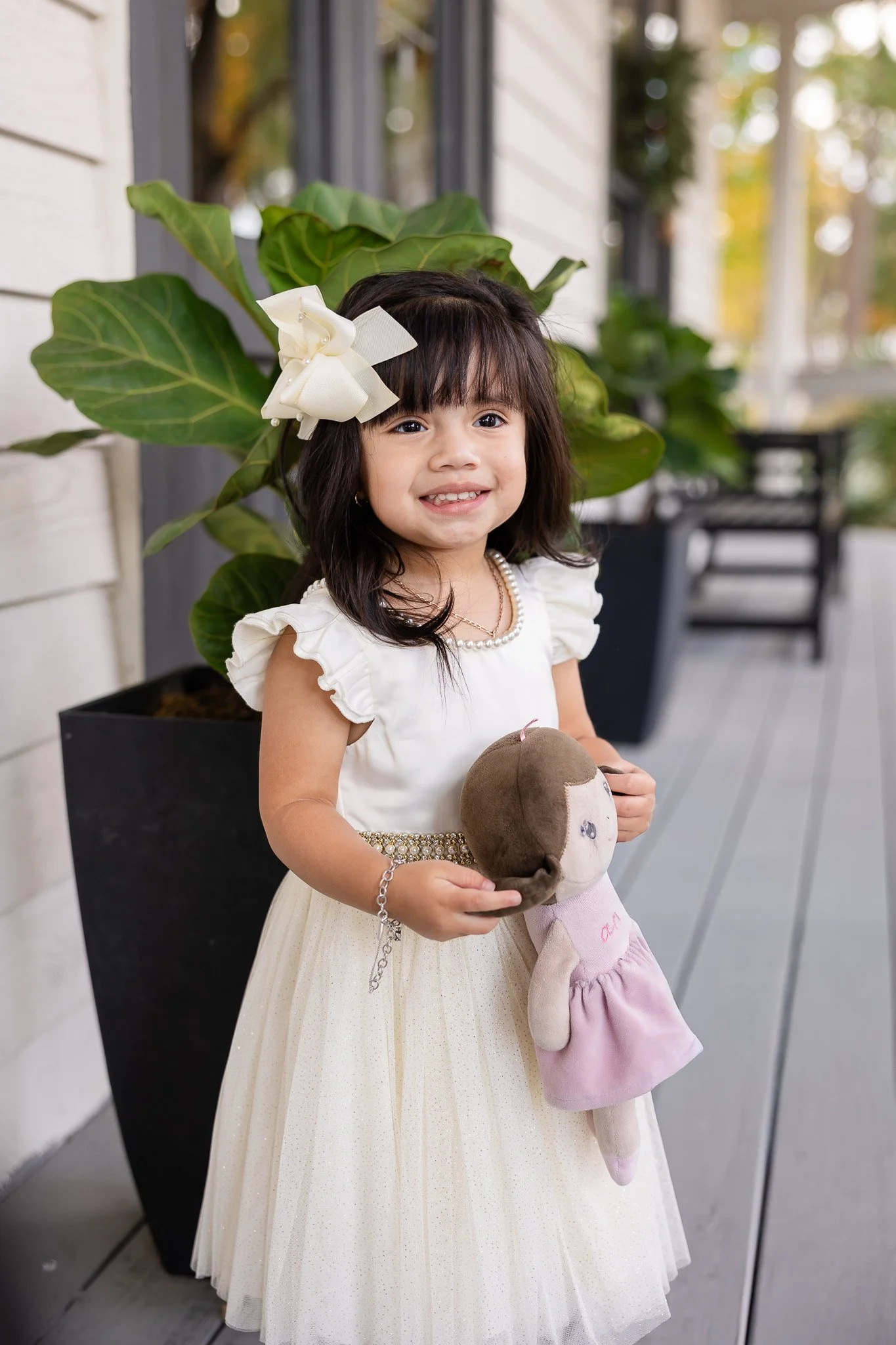 A young girl with dark hair and a white dress standing outdoors, holding a stuffed animal, with large green leaves and a potted plant in the background.