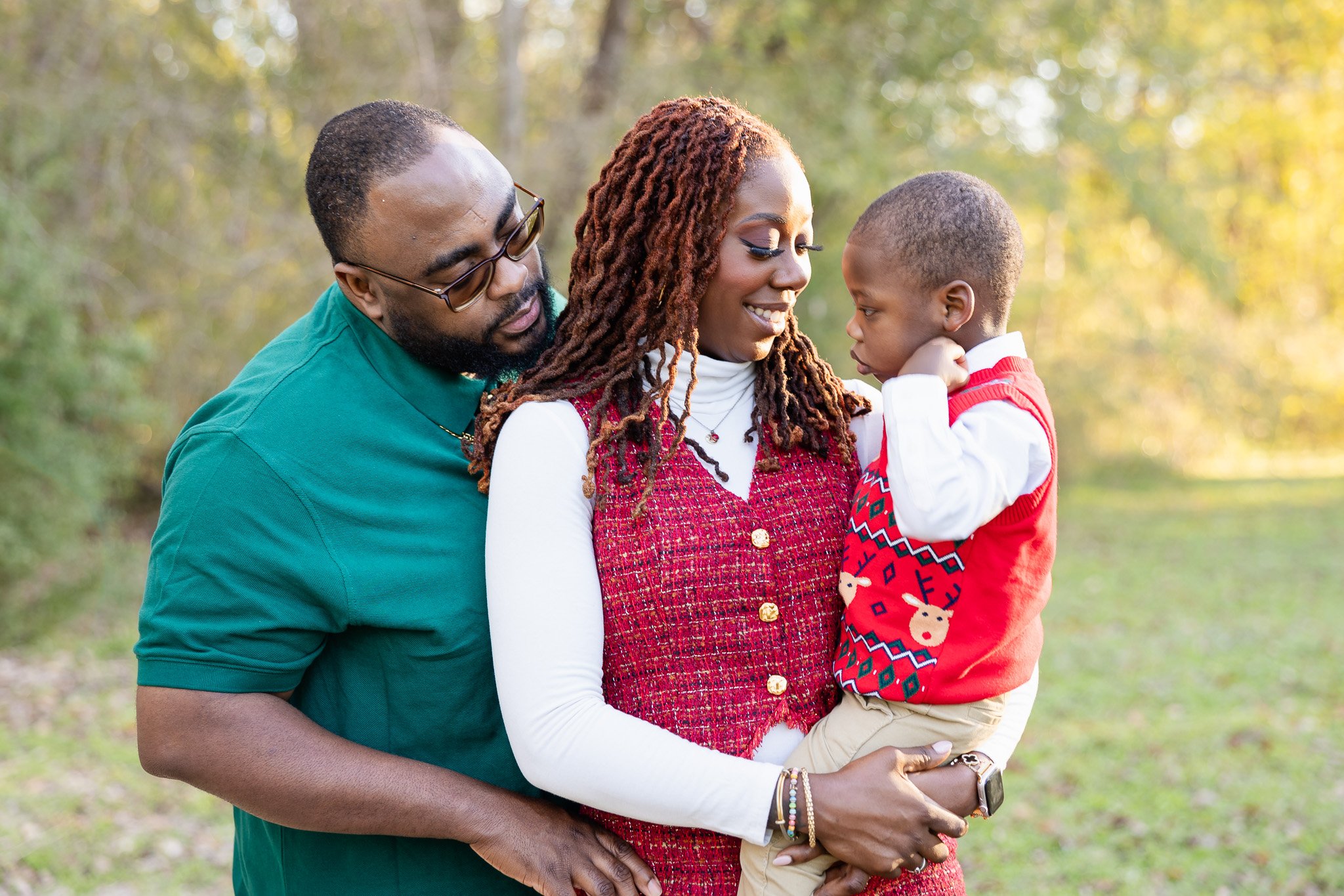 A family of three, outdoors in autumn, with the father on the left wearing glasses and a green shirt, the mother in the center wearing a red vest and white long sleeve shirt, holding their young son who is dressed in a Christmas sweater with reindeer pattern.