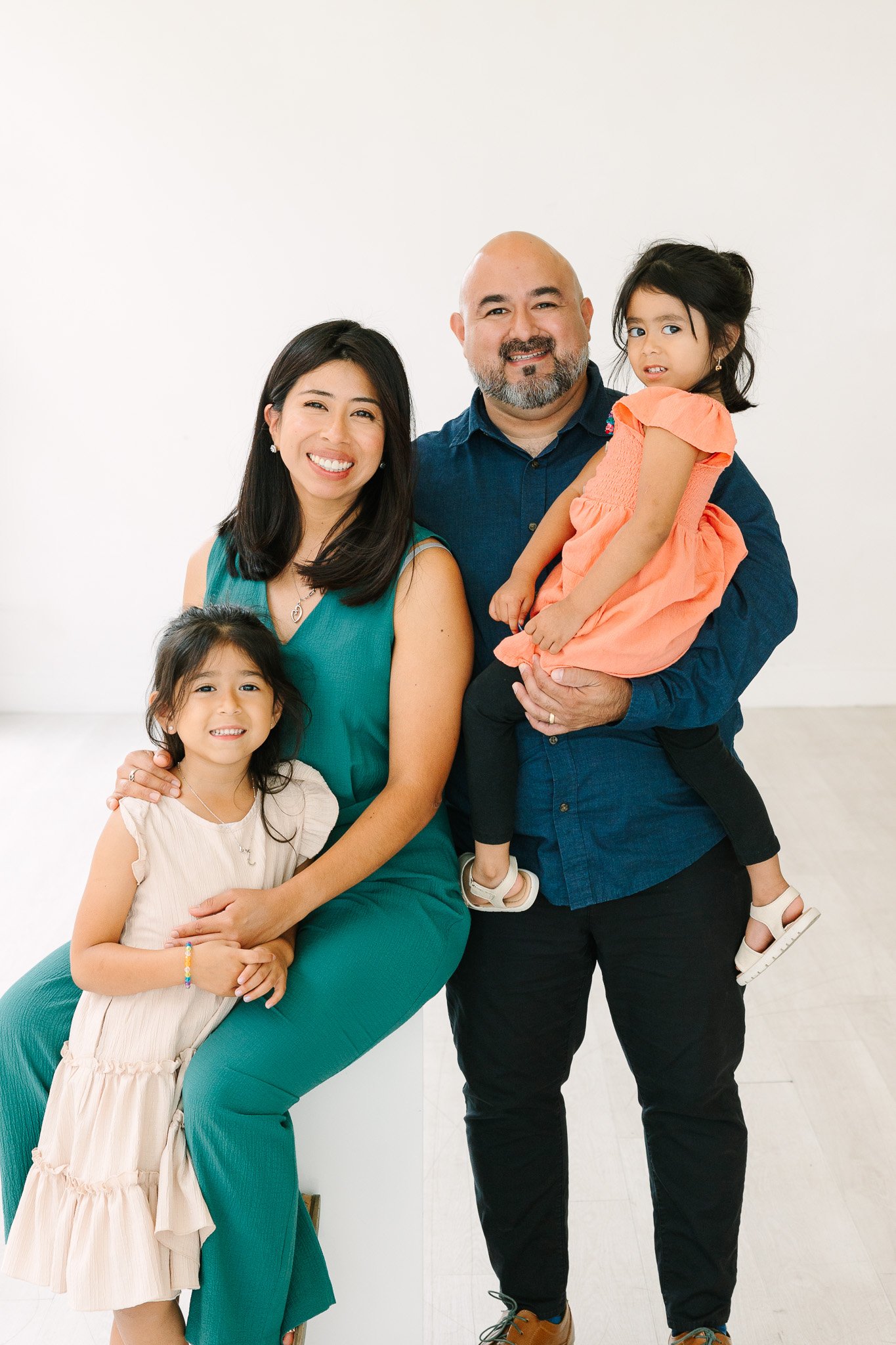 Family of four posing together with a man holding a young girl in an orange dress. The woman is sitting, smiling, wearing a teal dress. The girl in the beige dress is standing beside her, smiling. The man has a beard, is smiling, wearing a blue shirt, and holding the girl with dark hair and an orange dress.