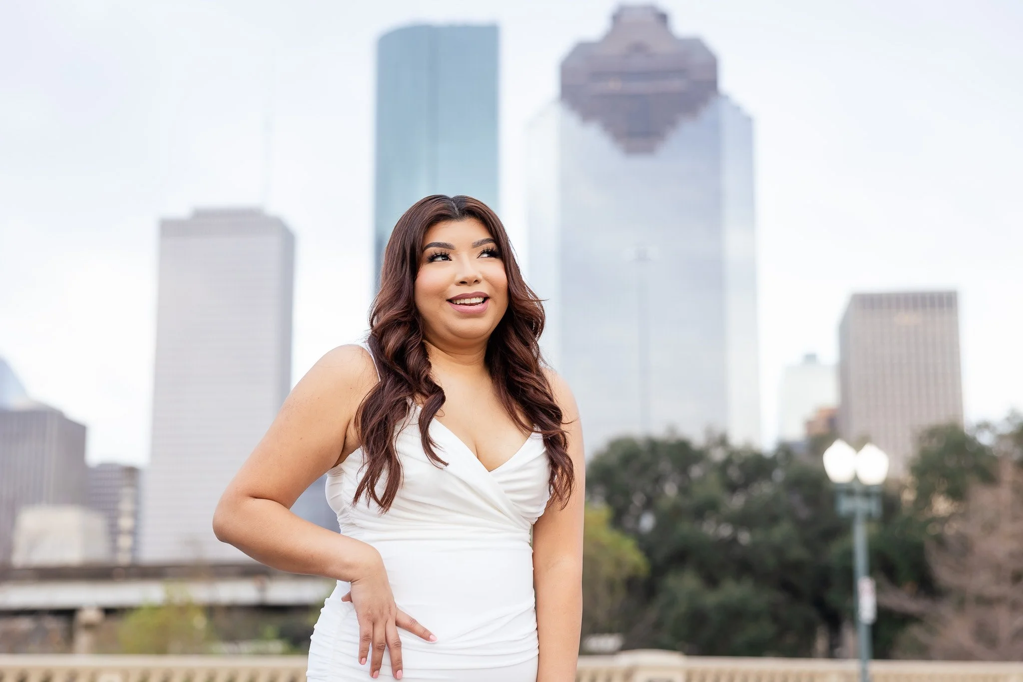 A woman with long, wavy brown hair and a white dress standing outdoors in front of tall city buildings, smiling softly.