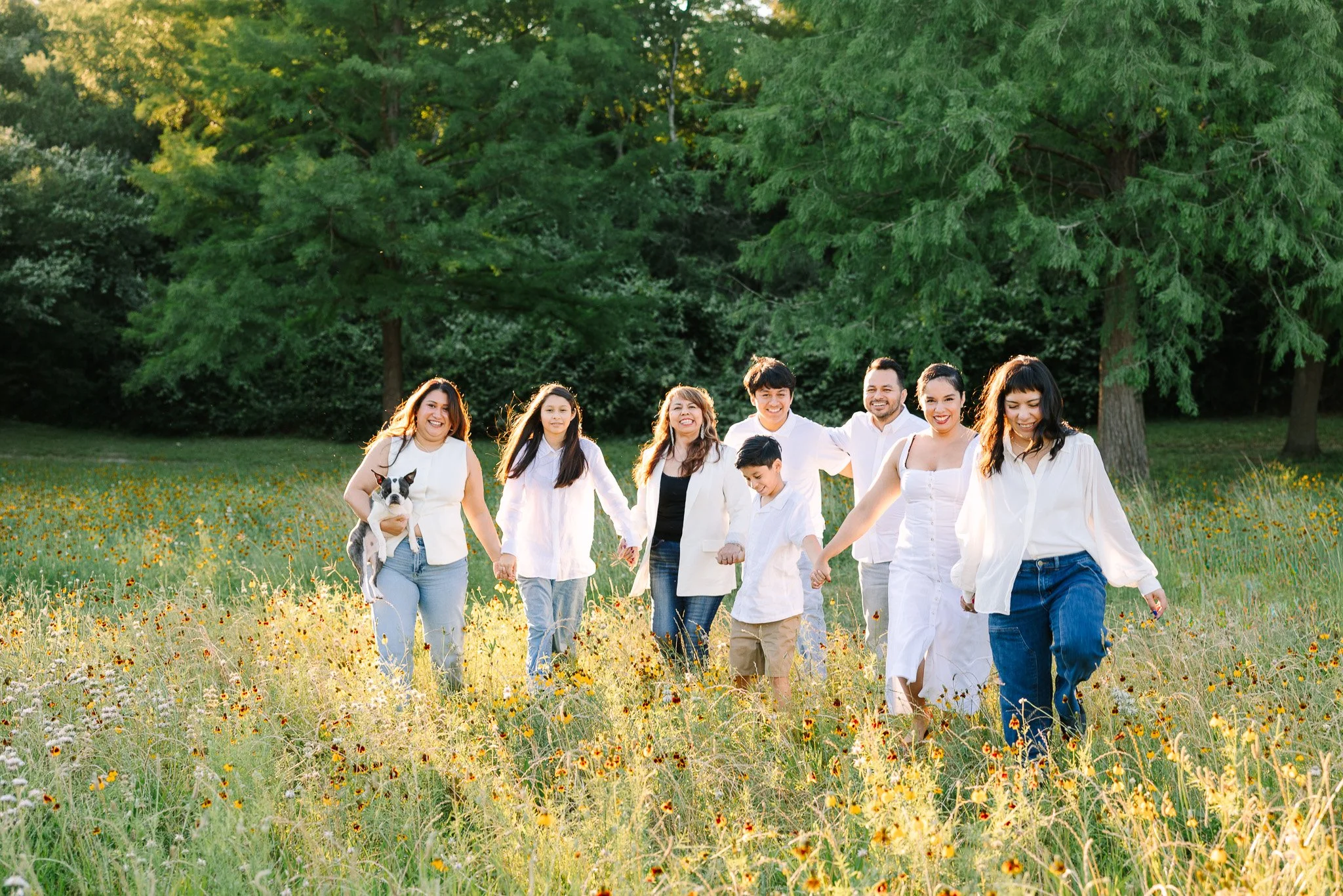A diverse group of people walking hand in hand through a field of yellow flowers in a park, surrounded by tall trees, during what appears to be late afternoon or early evening.