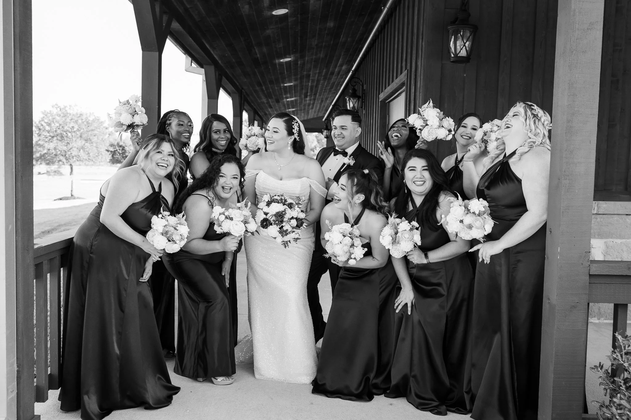 A black-and-white photograph of a joyful bride and her bridesmaids on her wedding day, standing in a rustic wooden structure with outdoor scenery in the background.