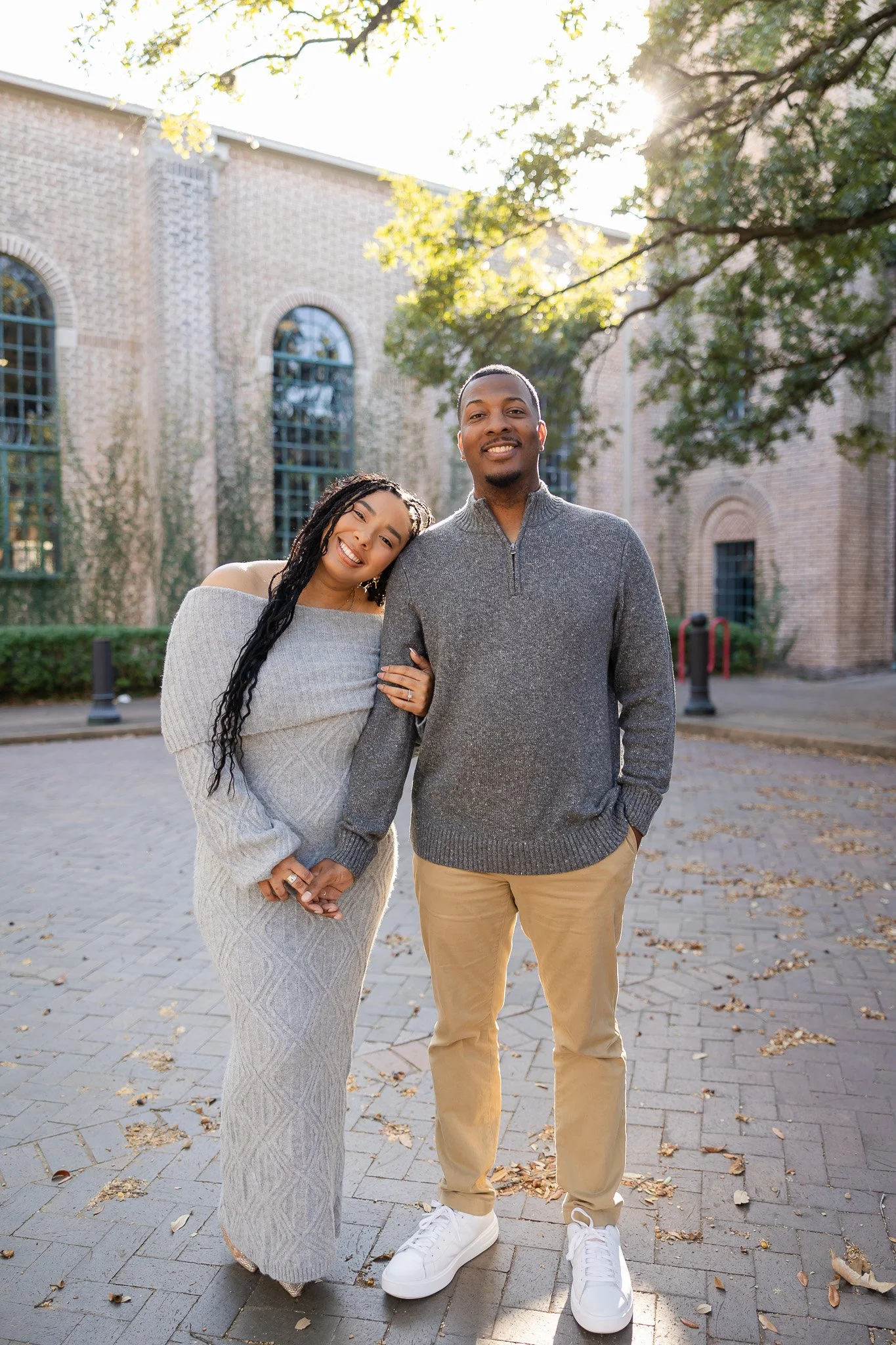 A smiling couple standing outdoors on a paved area with fallen leaves, holding hands, in front of a brick building with arched windows, trees with autumn foliage, and sunlight filtering through the branches.