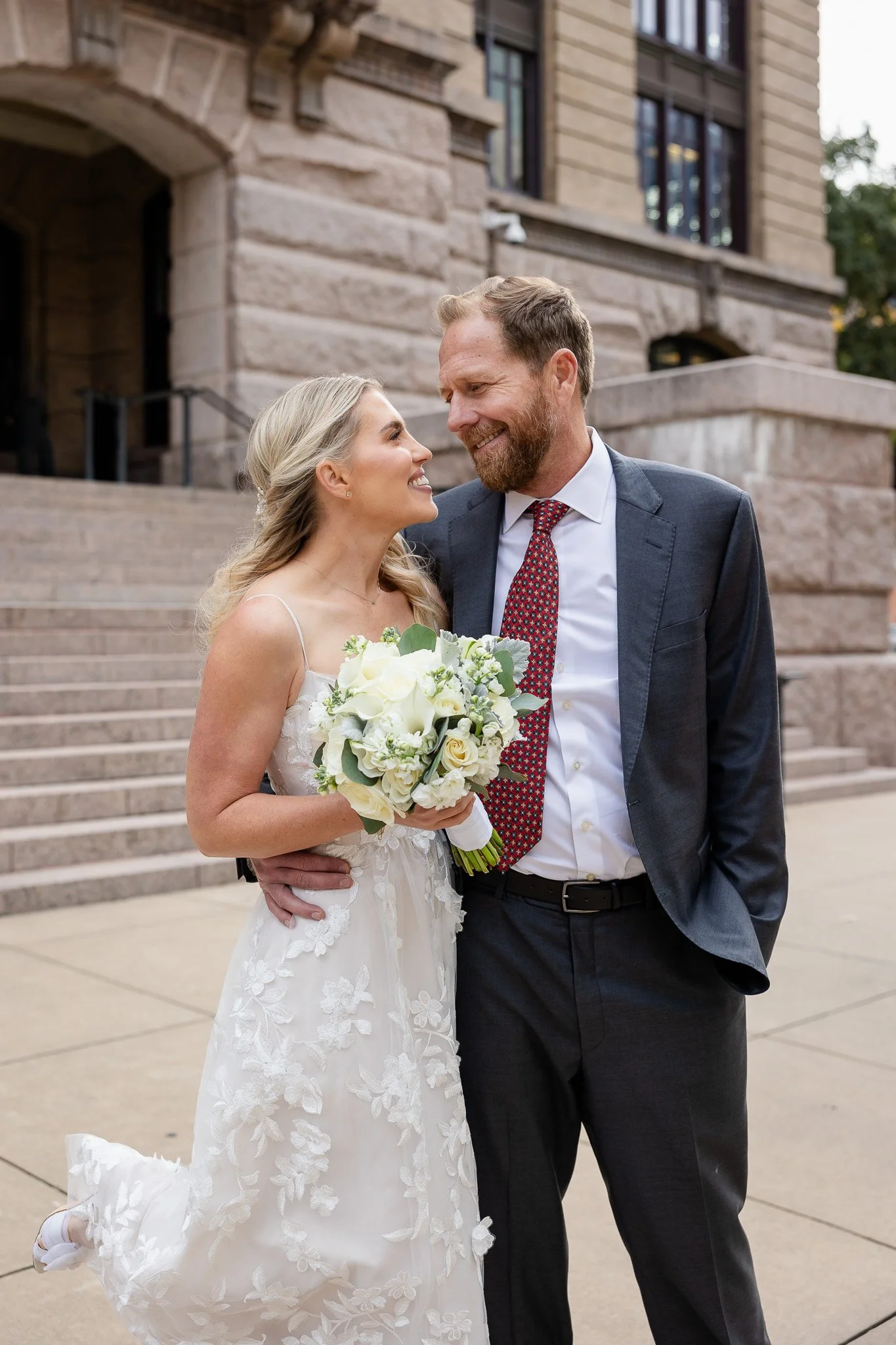 A bride and groom look at each other smiling outside a stone building, the bride holding a bouquet of white flowers, with steps behind them.