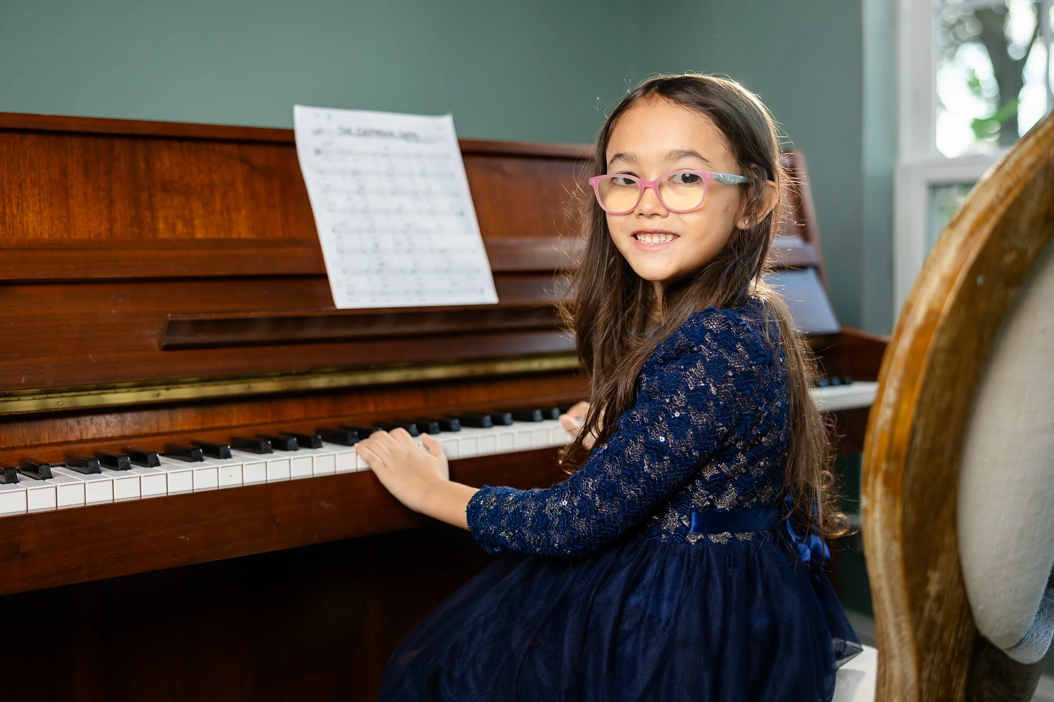 A young girl with long brown hair, pink glasses, and a blue lace dress sitting at a wooden piano, playing, in a room with a window and green walls.