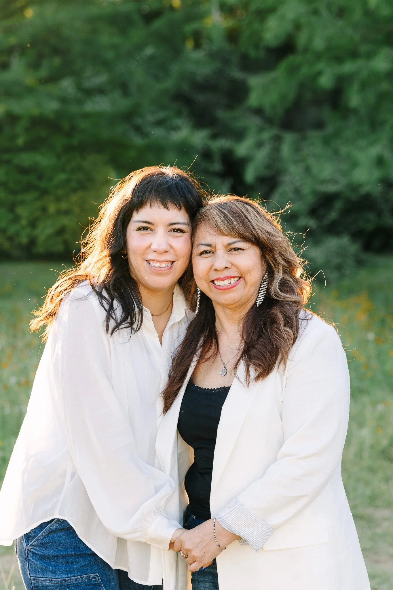 Two women standing outdoors close to each other and smiling, with a green blurred background.