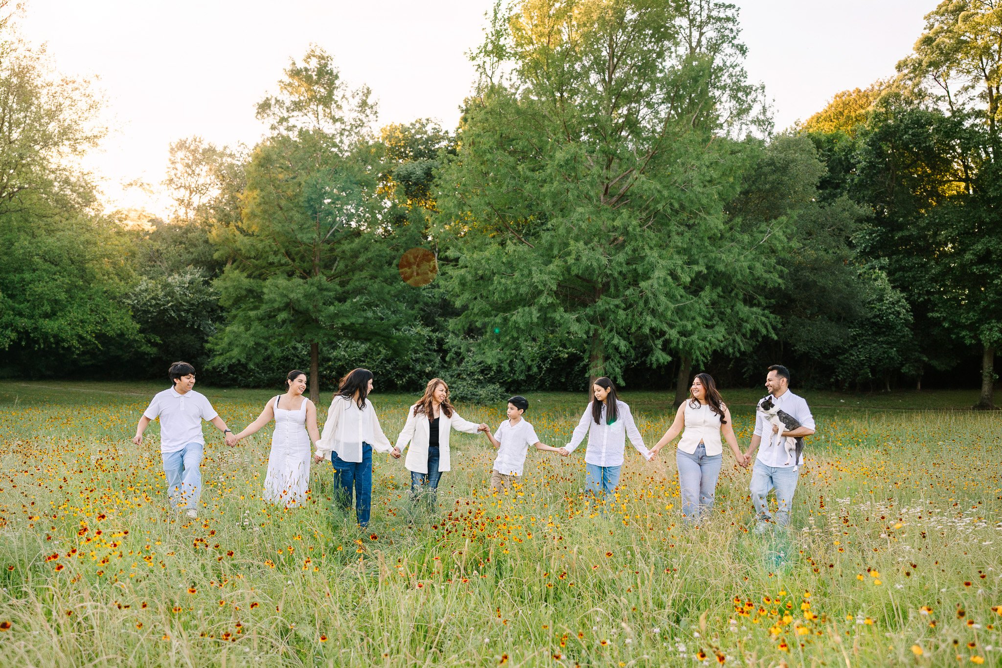Group of people holding hands in a circle in a grassy field with trees and a bright sky in the background.