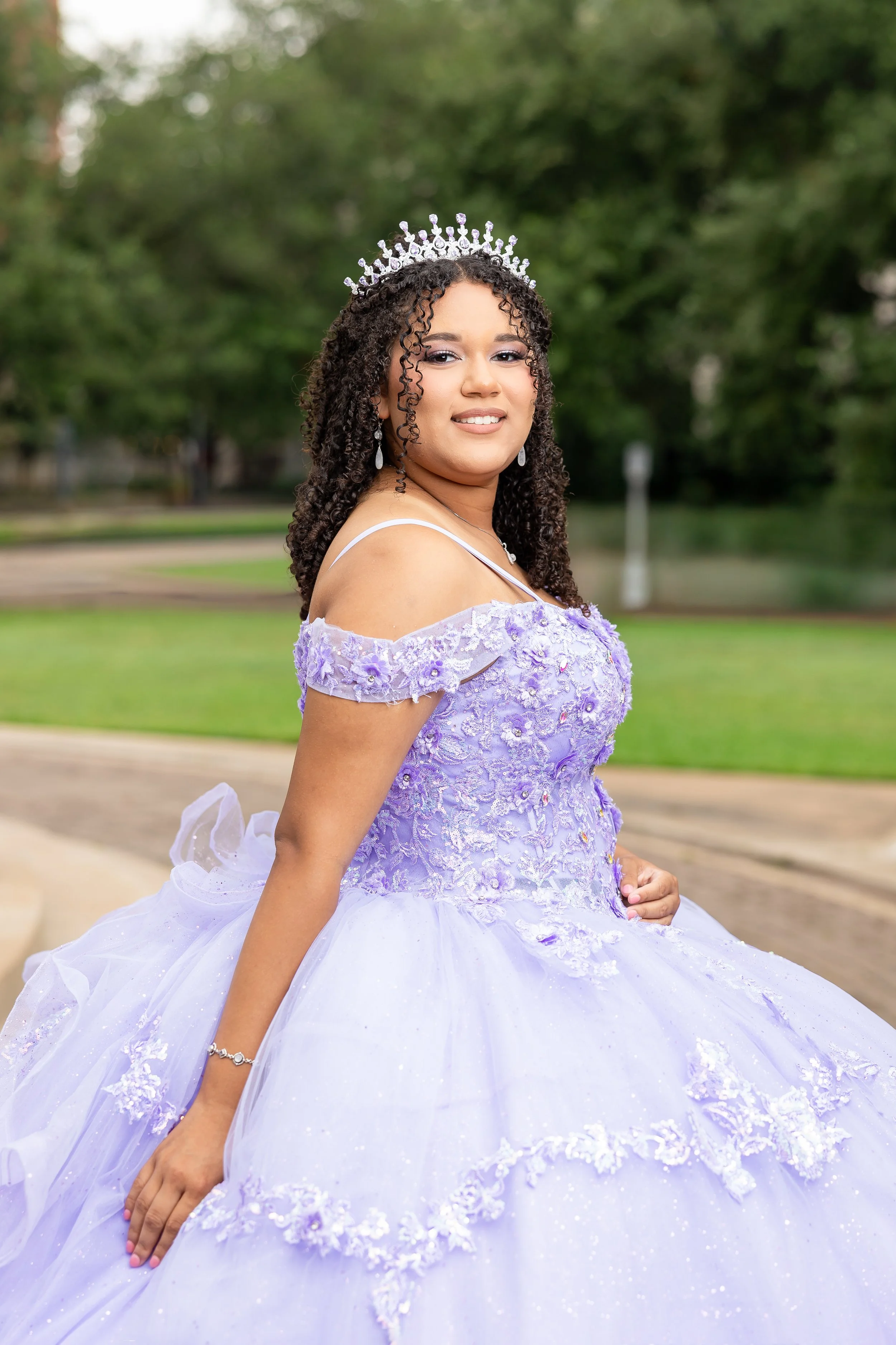 Young woman in a lavender and white quinceañera dress with off-the-shoulder ruffle sleeves, lace, and floral embroidery, wearing a crown and earrings, standing outdoors in a park with trees and grass in the background.