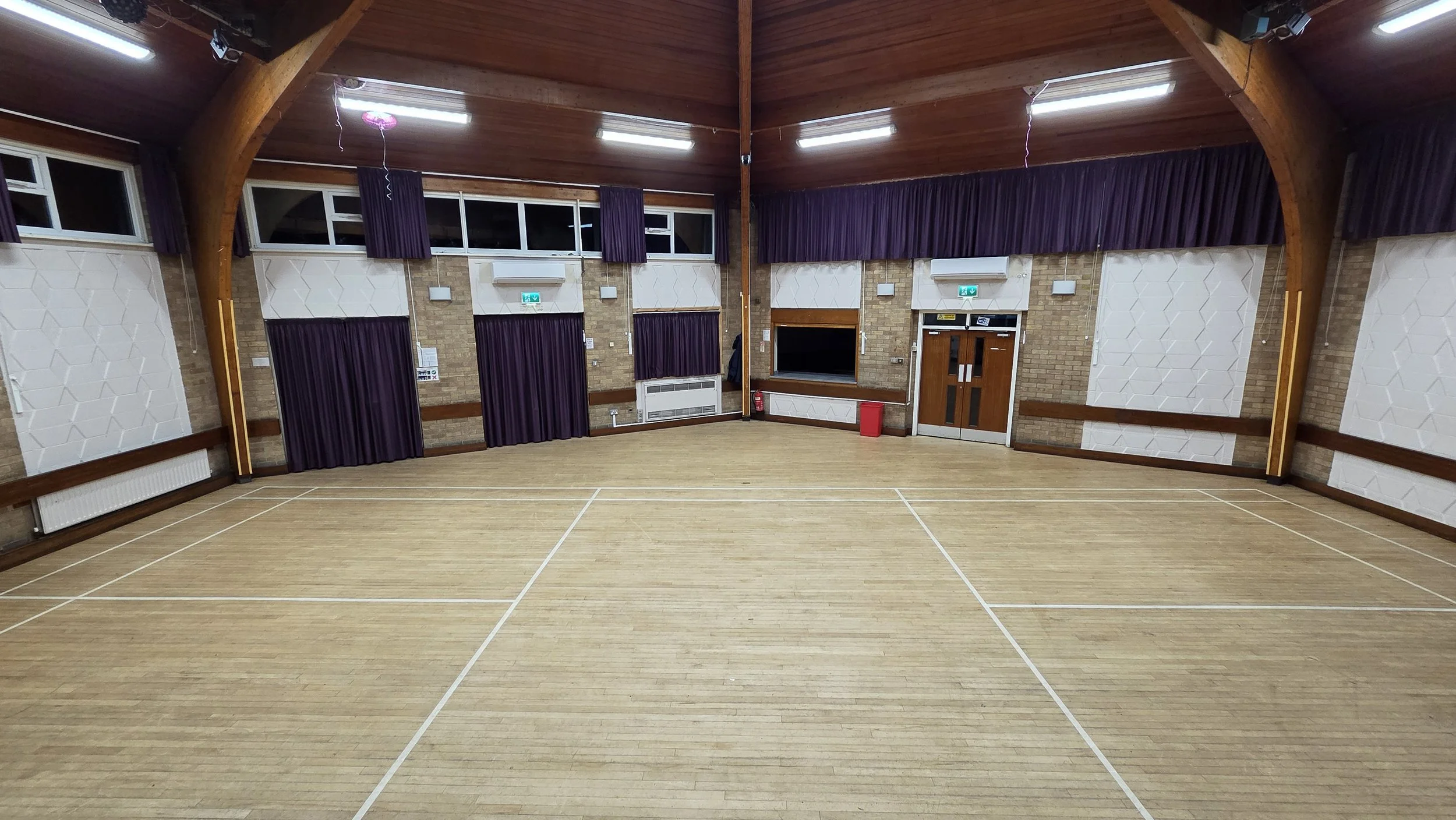 View of the Main Hall taken from the stage. The hall is slightly larger than a badminton court size. From left to right on the rear wall is the chair store, fire exit, storage room, kitchen servery hatch, main entrance doors from the lobby.