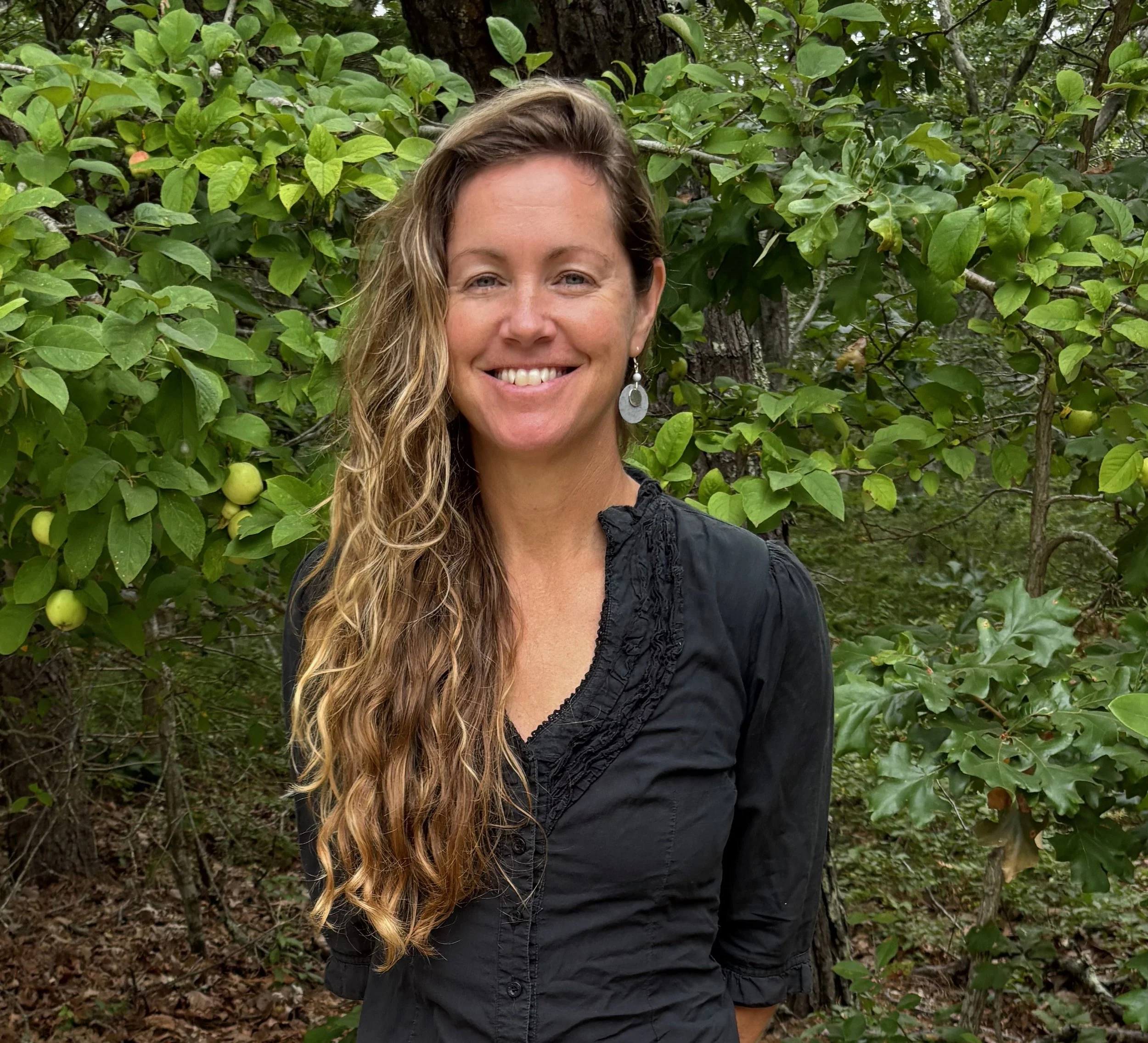 A woman with long brown hair smiling at the camera, wearing a gray shirt and a red top underneath against a brown background.