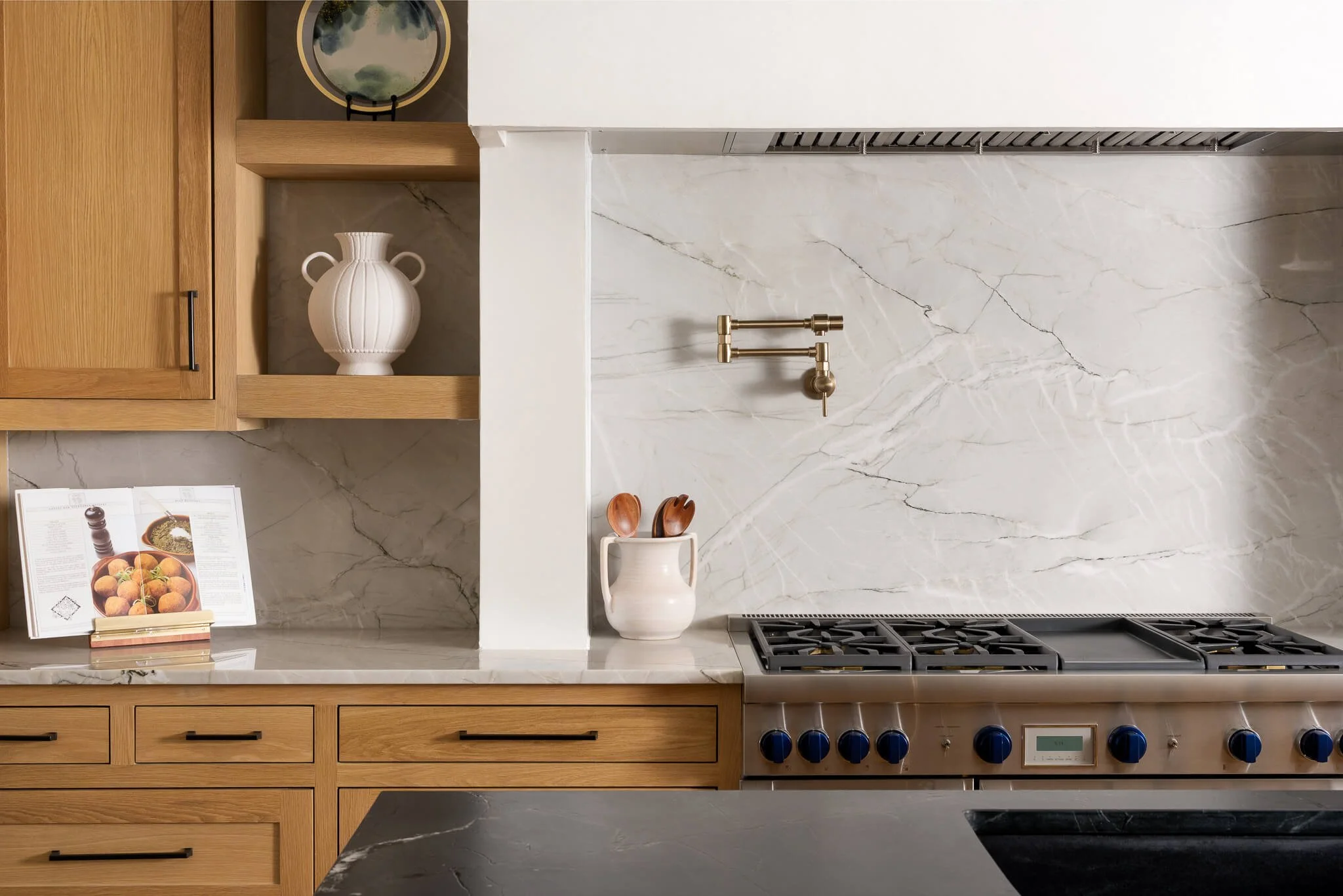 Kitchen with wooden cabinets, marble countertop, open shelf with white vase, stove, open cookbook, and utensils in a mug.