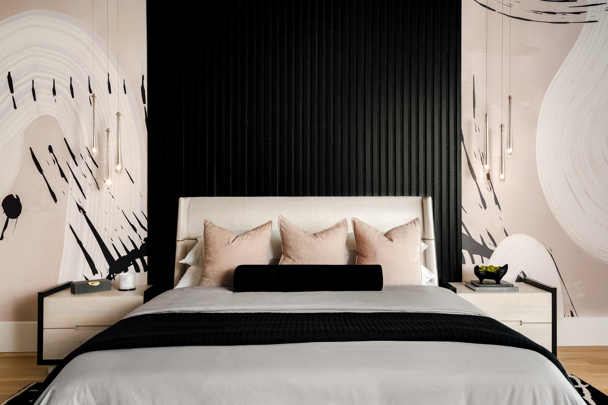 Modern bedroom featuring a beige upholstered bed with pink pillows, black and white abstract wall art, pendant lights on either side, a black nightstand with decorative items, and a black and white rug.