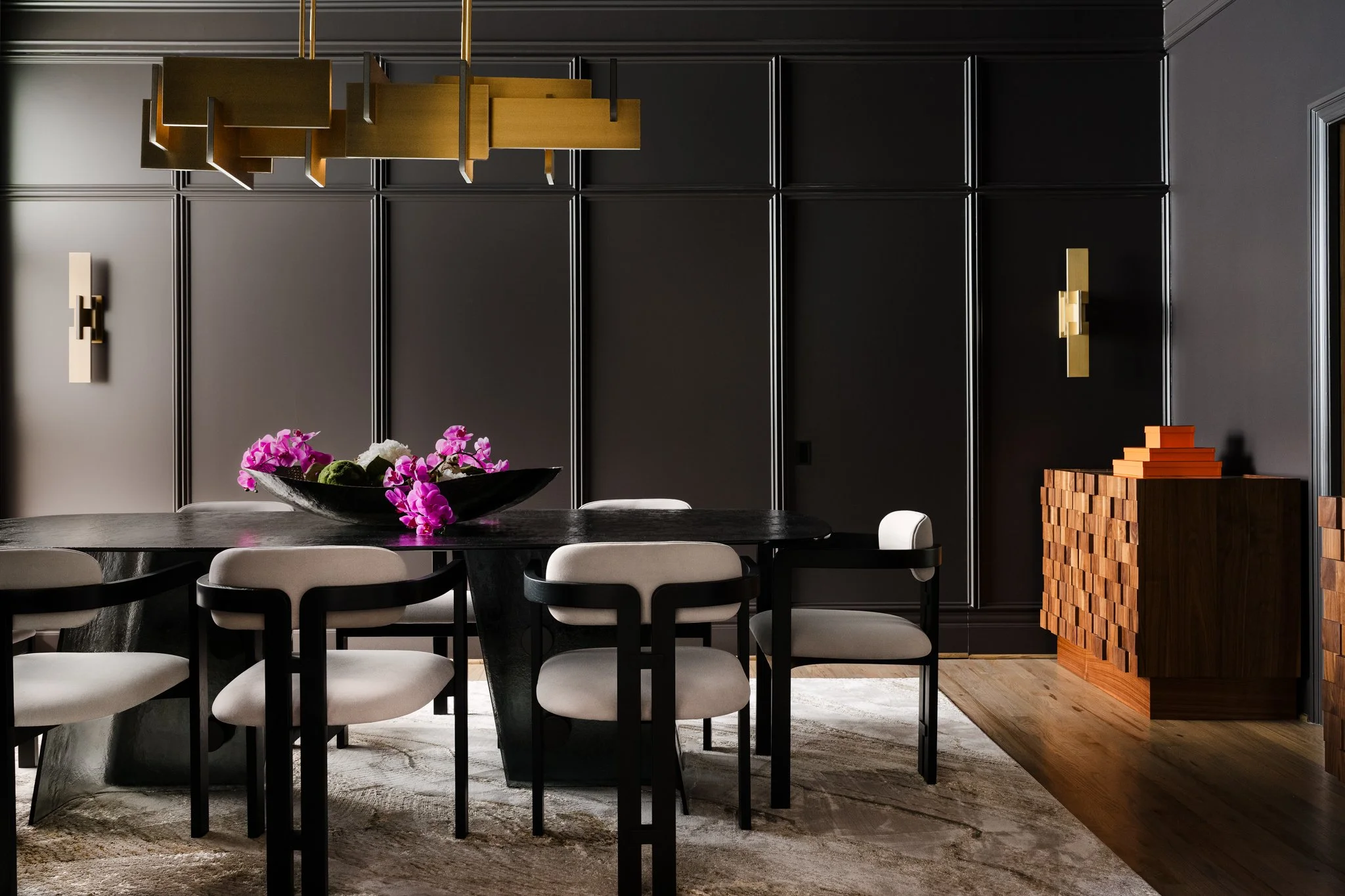 Elegant dining room with black walls, white chairs surrounding a dark oval table, pink orchid centerpiece, and wooden accents including sideboards and wall sconces.