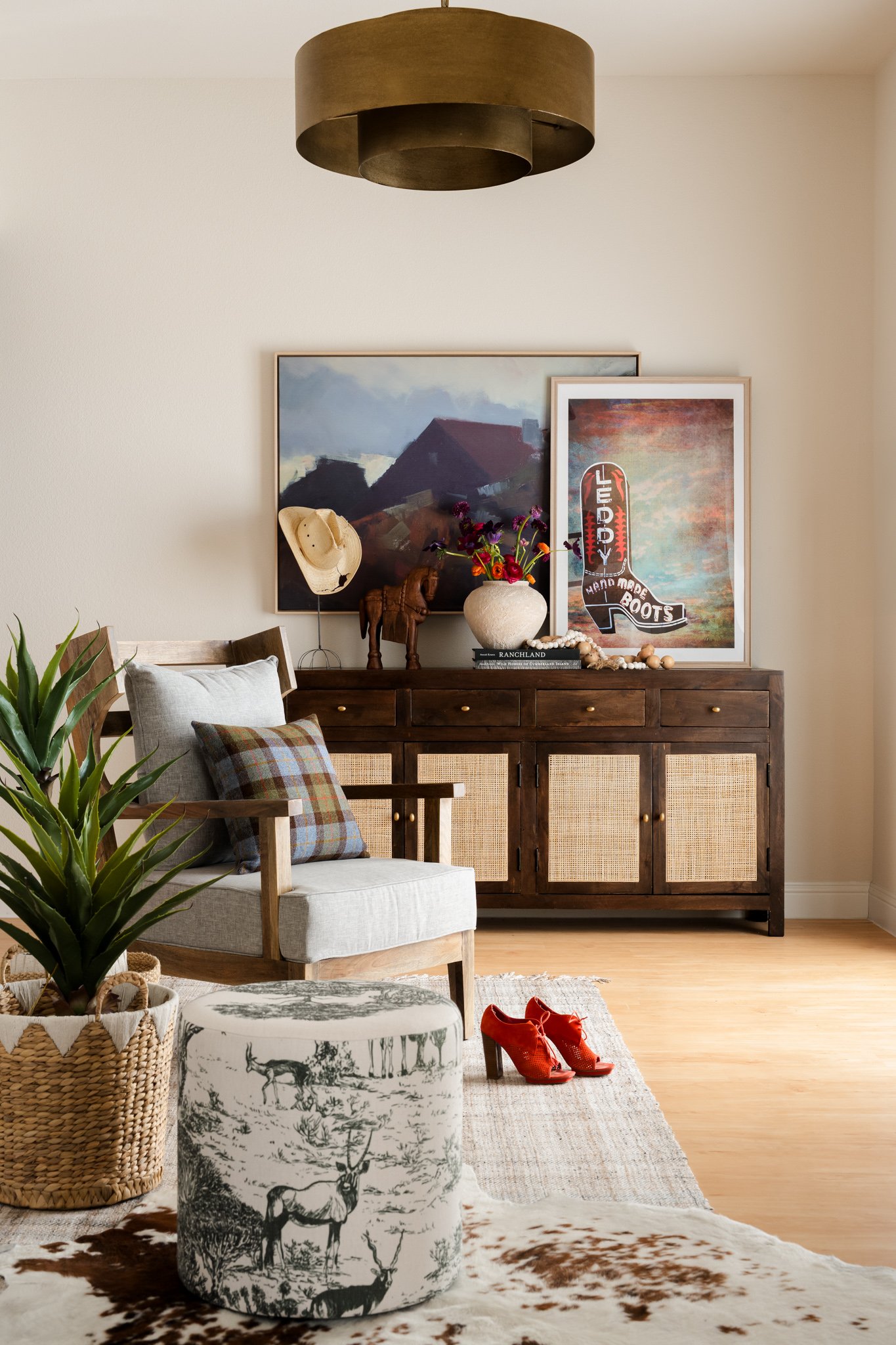 Living room with a wooden sideboard, framed artworks, decorative items including a hat, a horse sculpture, a vase with flowers, and a stack of books; armchair with plaid pillow, potted plant, a cowhide-patterned ottoman, and a red pair of shoes; ceiling light fixture; wooden flooring, beige walls.