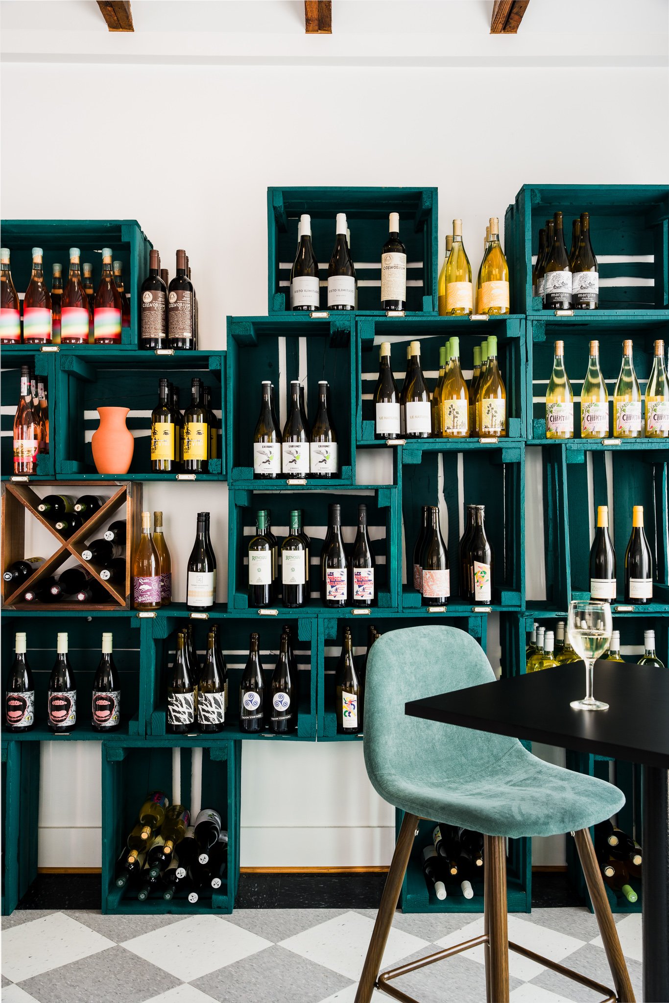 Wine bottles arranged in teal wooden shelves in a wine shop or tasting room, with a table and green upholstered chair with wooden legs in the foreground.