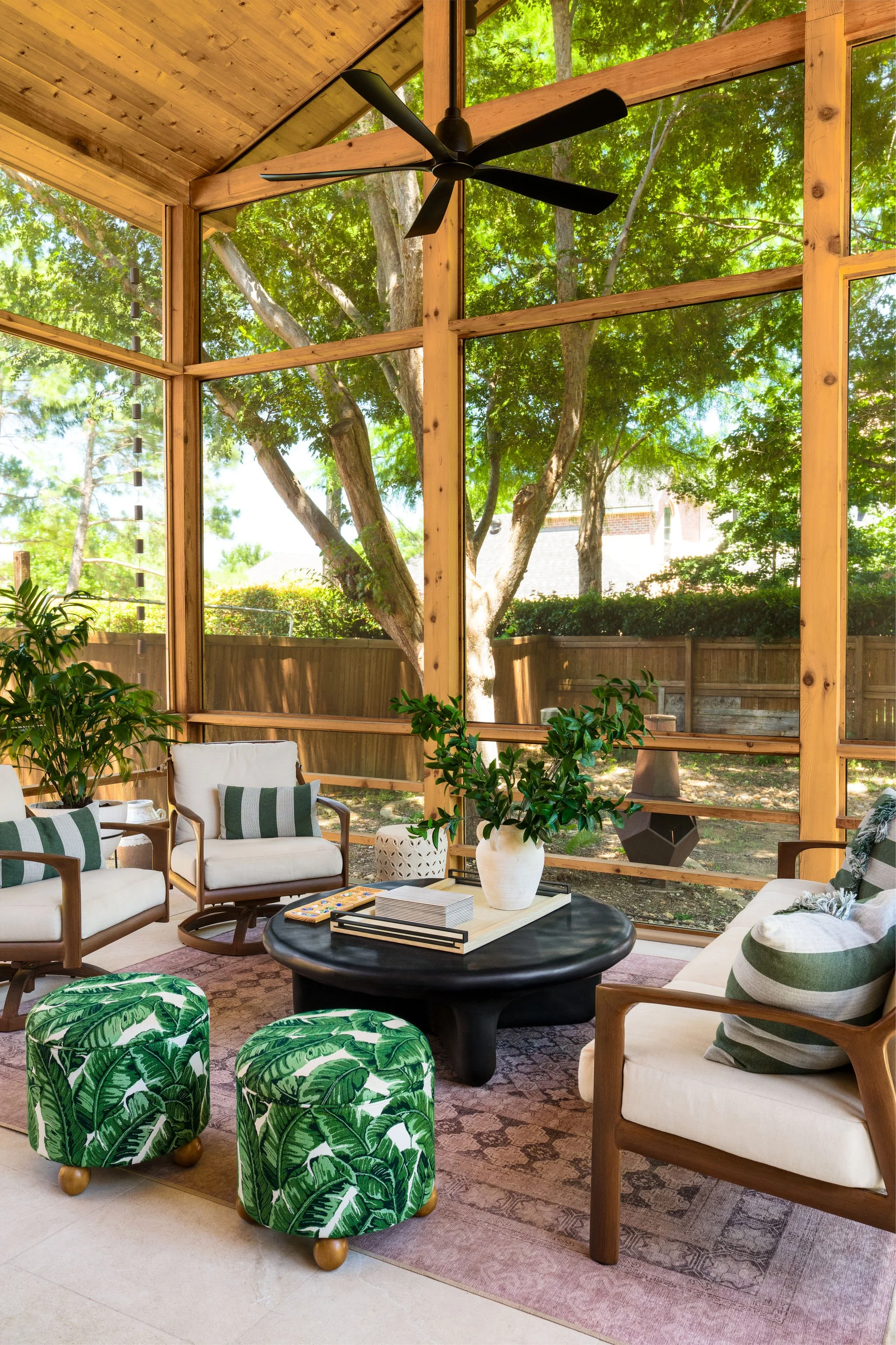 A bright and airy screened-in patio with a travertine dining table, black sculptural chairs, lush plants, and tropical ottomans—perfectly styled