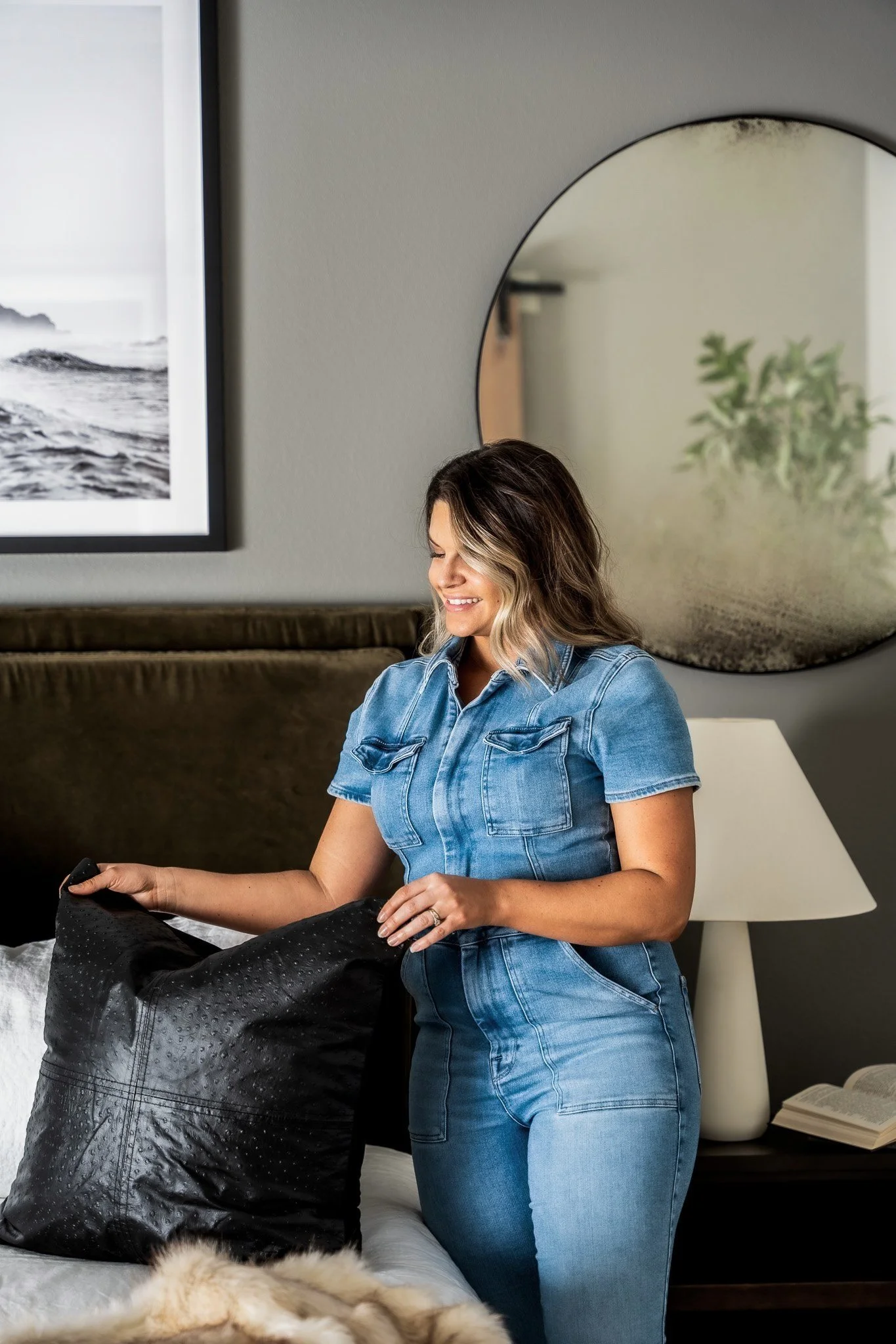 A woman in a denim jump suit smiling and adjusting a black pillow on a bed in a modern bedroom.