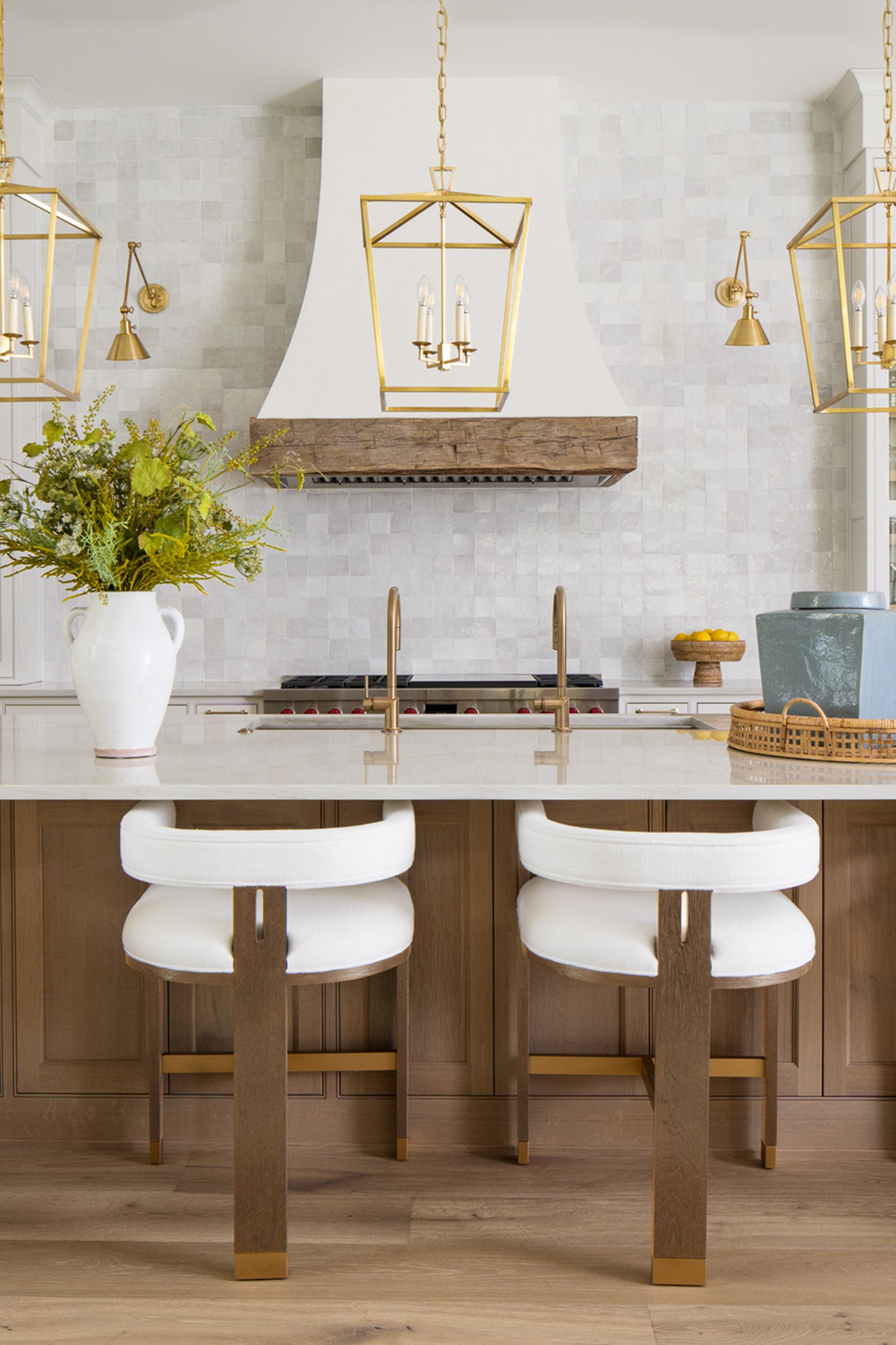 A kitchen island with a white countertop, two white chairs with wooden legs, a large white vase with green and yellow flowers, and a fireplace with a wooden beam and brass fixtures. There are golden chandeliers and wall sconces, and a bowl of lemons on the right side.
