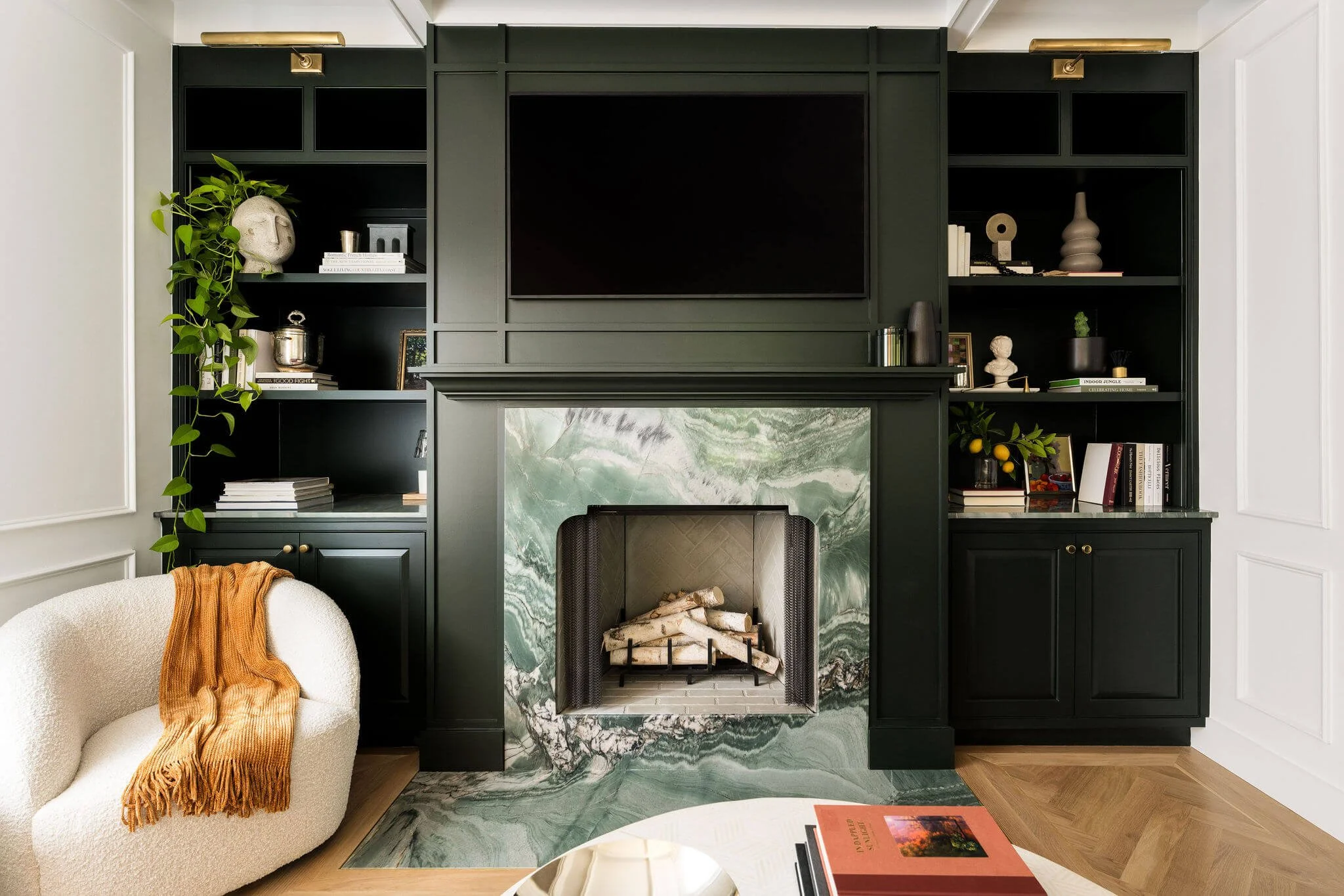 A living room with a dark green built-in bookshelf and a marble fireplace with a TV mounted above.
- Left side of the fireplace has books, decorative objects, and a plant.
- Right side has books, vases, and a small sculpture.
- Beige armchair with a 