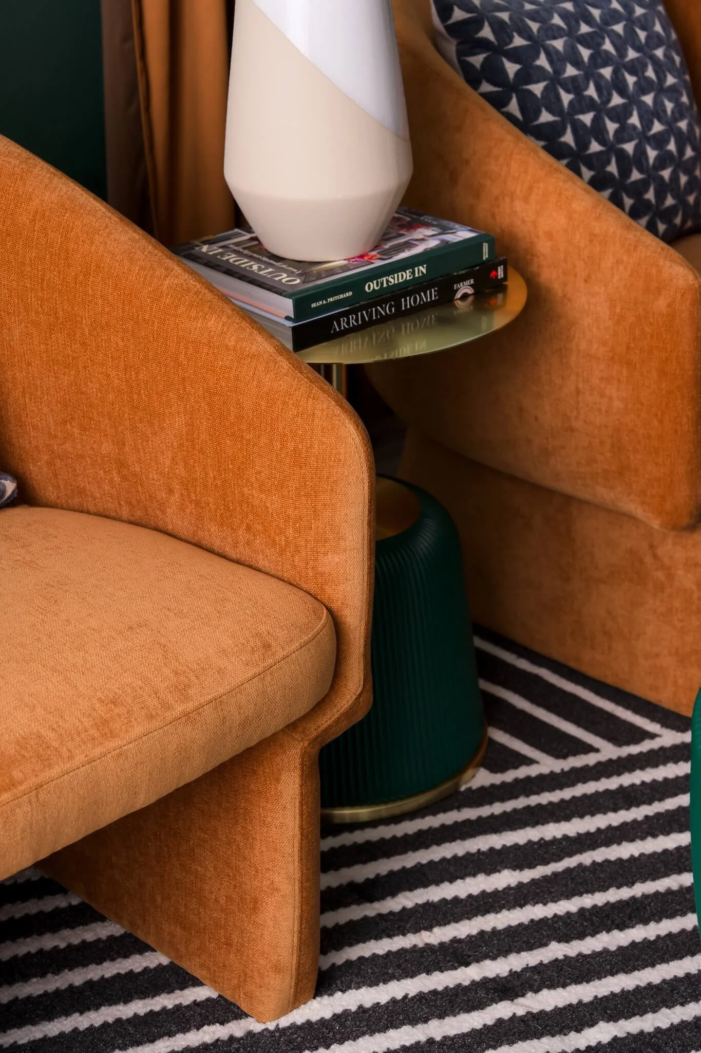 Close-up of a room corner with an orange velvet armchair, a black and white striped rug, and a small side table holding a white geometric vase and a stack of books.