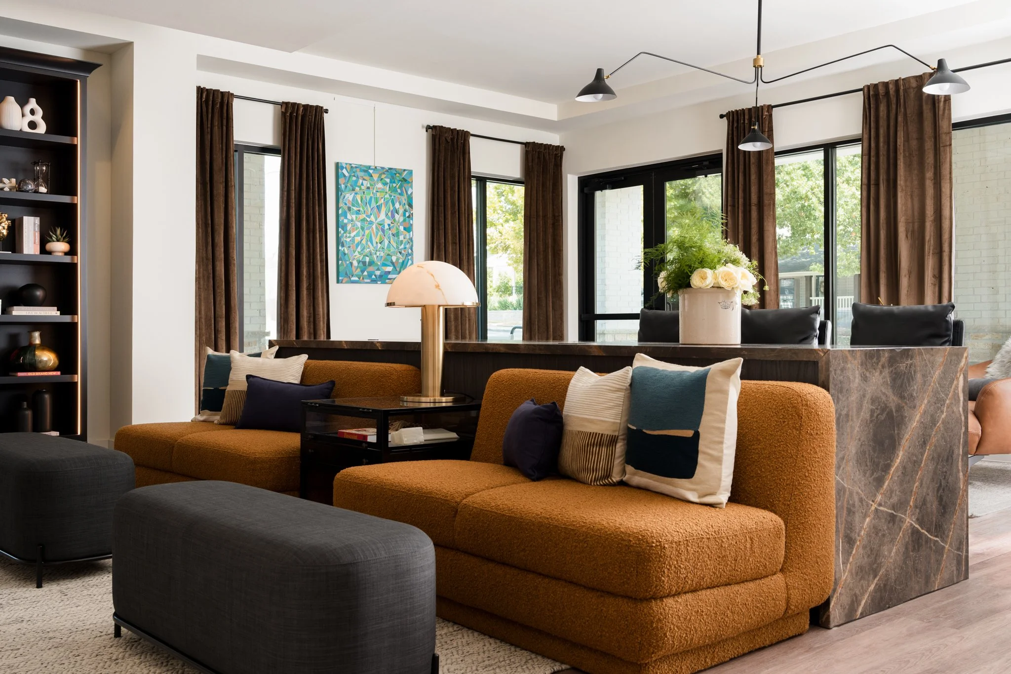 Living room with brown curtains, large windows, contemporary black pendant lighting, a colorful abstract painting, a white flower arrangement on a marble and wood table, and a mix of orange, black, and striped pillows on sofa and chairs.