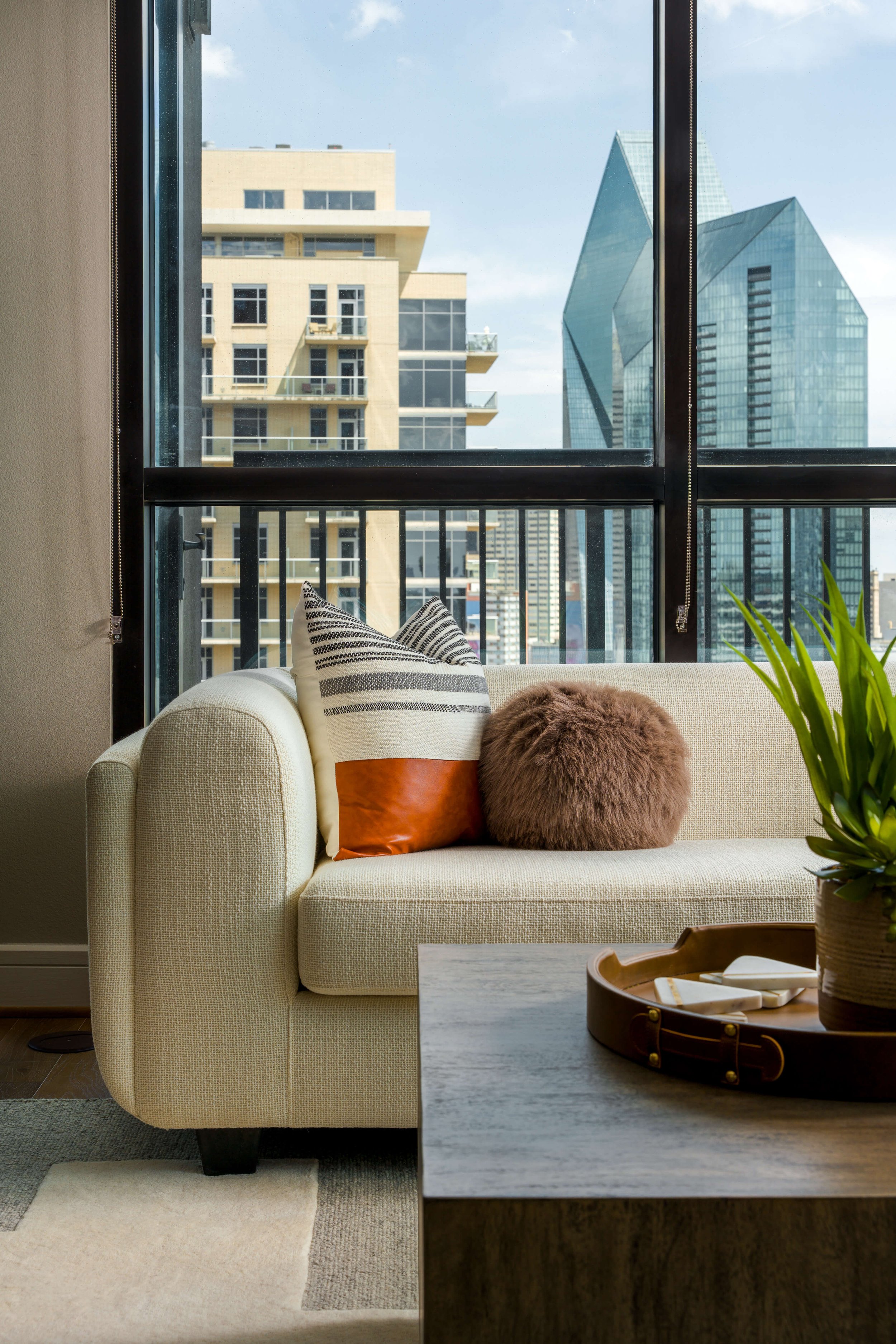 This modern high rise apartment designed by Travis Pernell Interiors features views in every direction, a cream colored sofa, textured orange and tan pillows, and a cognac leather coffee table tray. 
