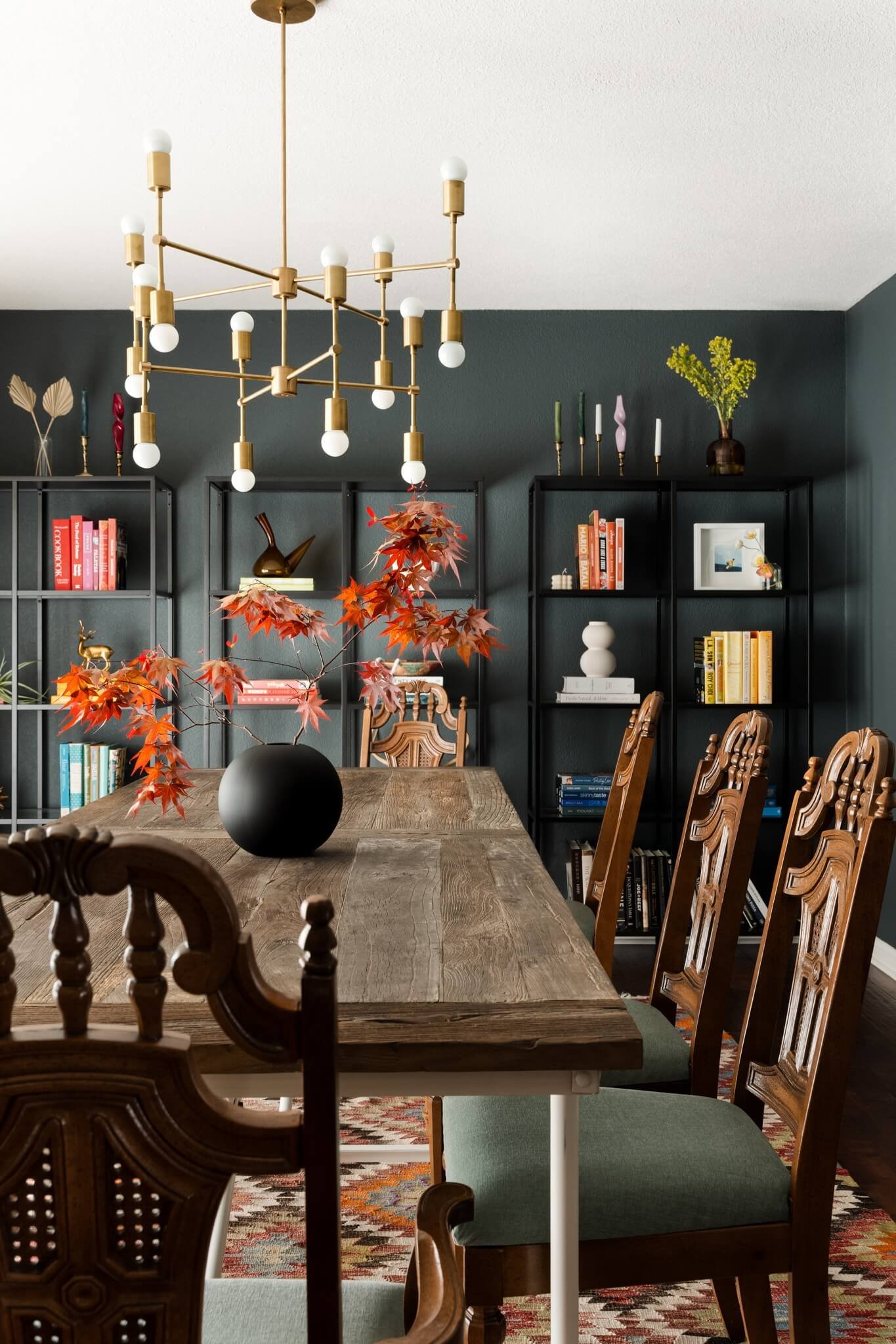 A dining room with a wooden dining table, surrounded by wooden chairs with carved backs and green cushions. A black vase with red and orange autumn leaves sits on the table. Behind the table are black bookshelves with various books, decorative object