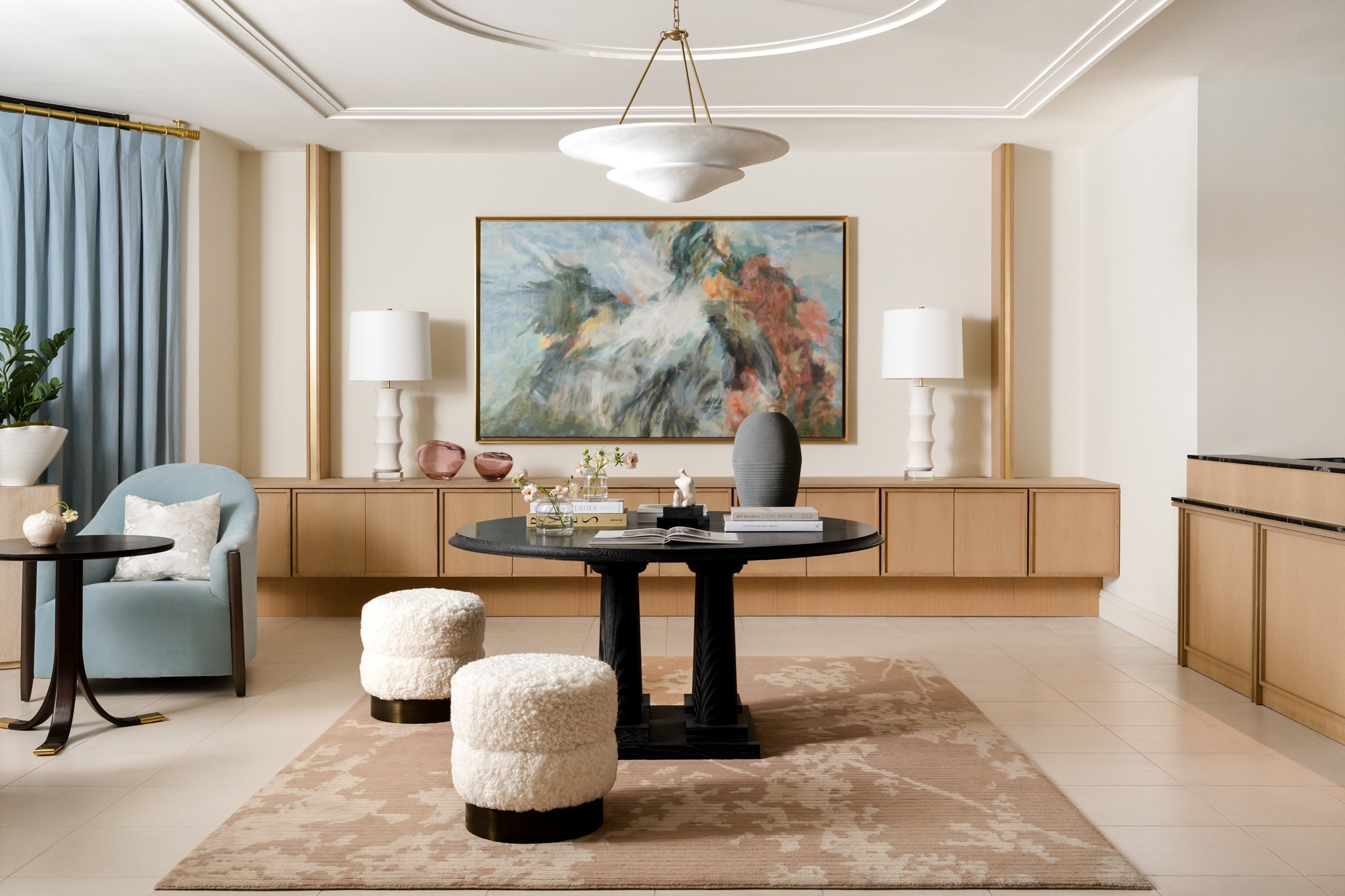 A modern living room with a light-colored wall, wooden sideboard, abstract painting, white lamps, a round black table, upholstered chairs, and decorative items including vases and books. A beige area rug covers the floor.