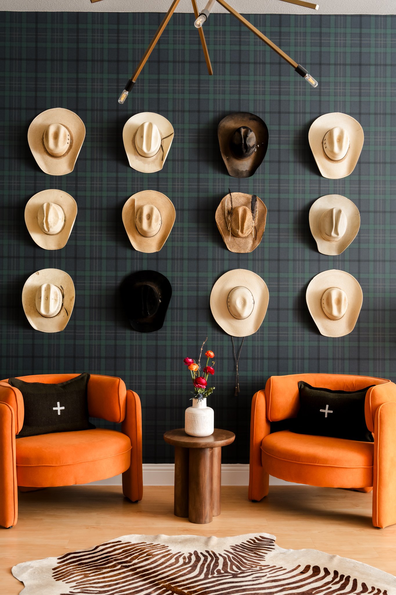 Living room with a navy blue plaid accent wall decorated with multiple cowboy hats arranged in a grid pattern. Two orange armchairs with black pillows featuring white plus signs are positioned on either side of a small round wooden table with a white