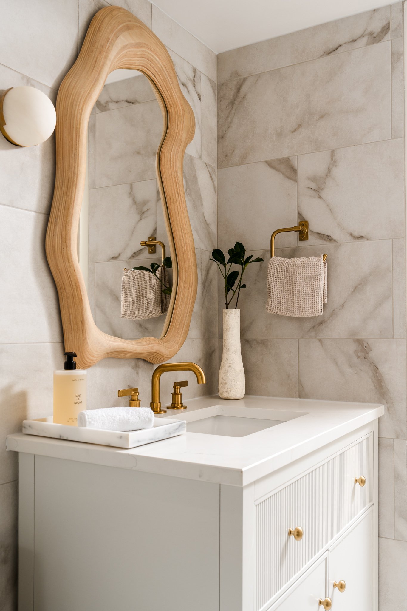 Bathroom vanity with a white sink, gold fixtures, a soap dispenser, a rolled towel, a potted plant in a tall vase, a wooden-framed mirror, a wall sconce, and beige marble tiles on the wall.