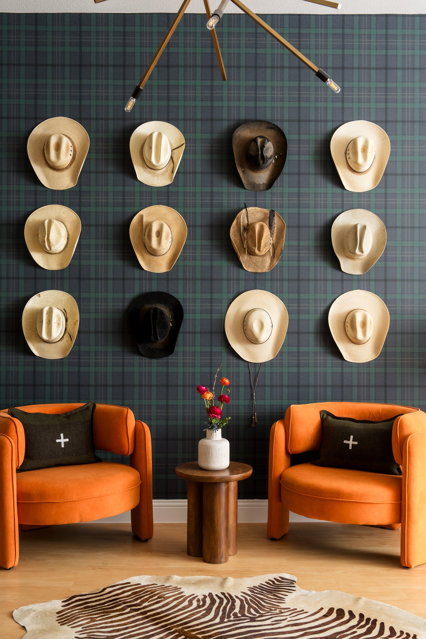 Interior of a room with a dark plaid wallpaper decorated with twelve cowboy hats arranged in four rows. Two orange armchairs with black pillows, a small wooden table with a white vase and pink flowers, and a zebra-print rug are also visible.