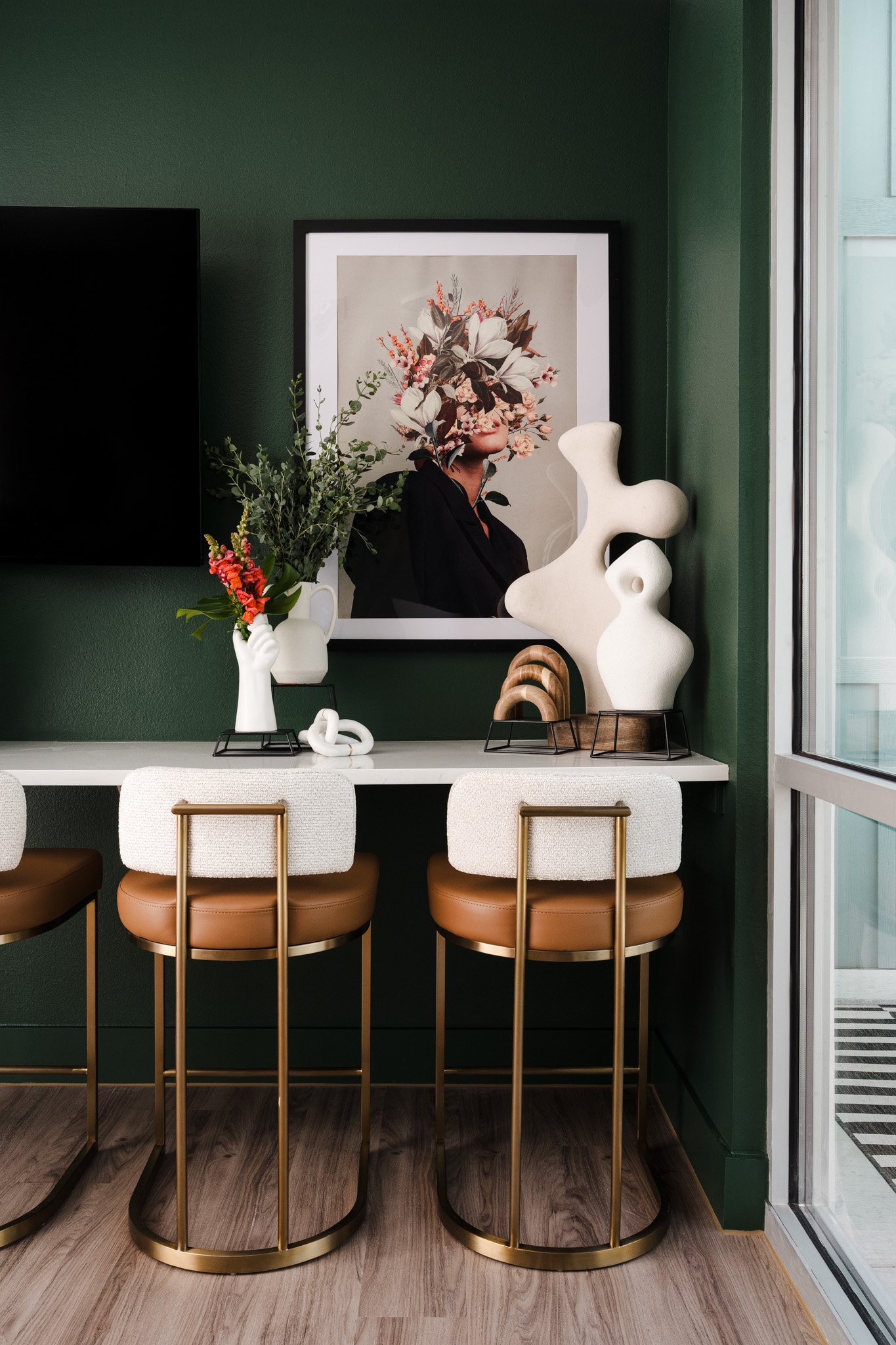 Interior of a modern dining or kitchen area with a white countertop, three chairs with beige padded seats and gold metal frames, a large green wall with a framed floral artwork, decorative vases with flowers and abstract sculptures, and a sliding gla